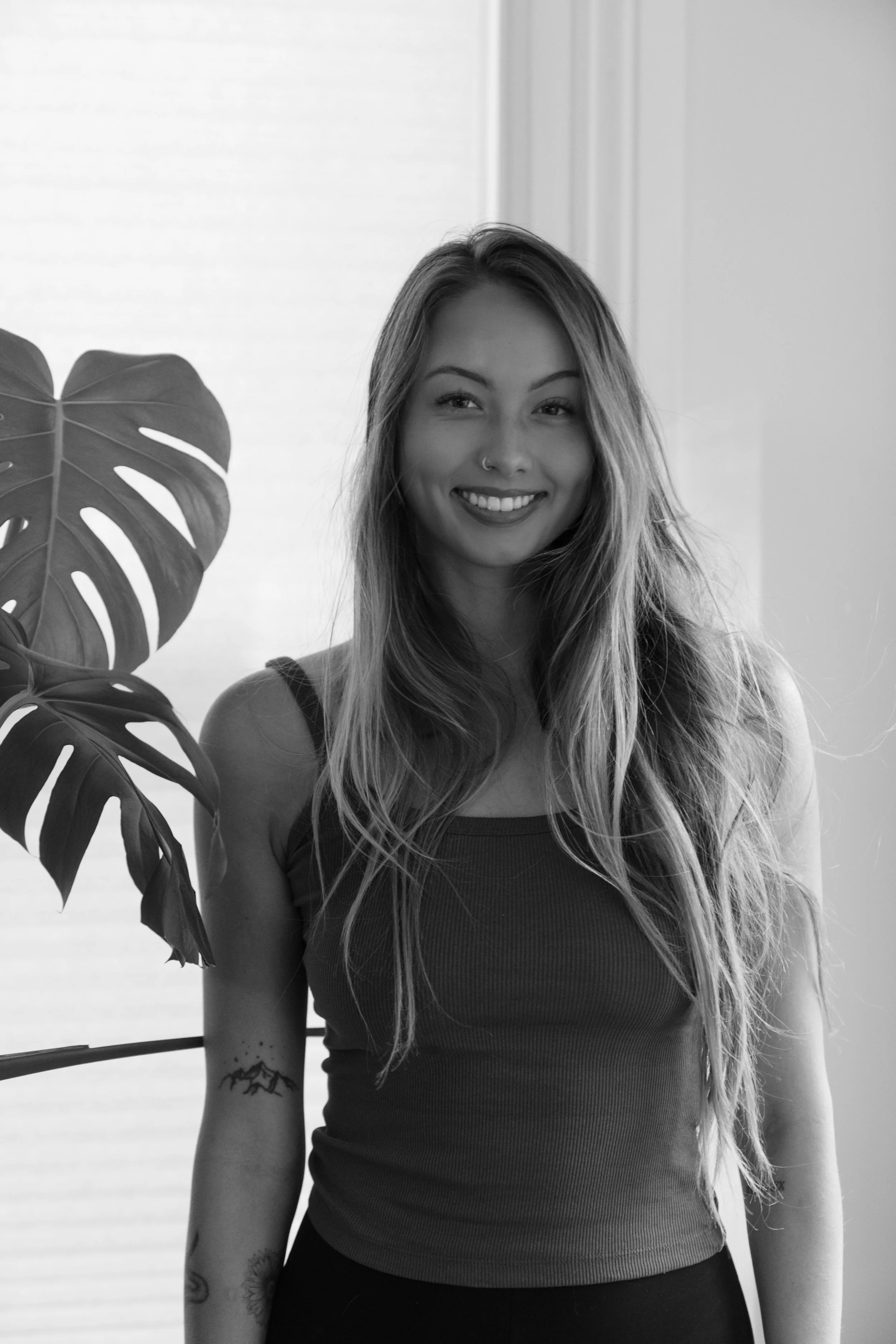 A black and white image of a pilates instructor standing next to a large plant and posing for a headshot at a photo studio in Providence, Rhode Island.