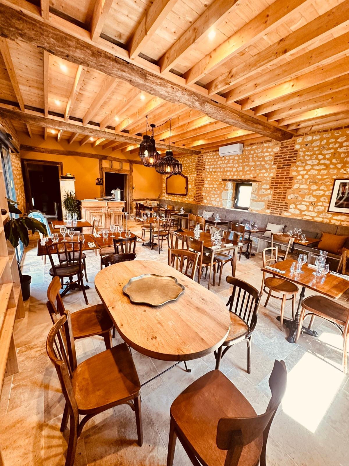 Une salle à manger chaleureuse avec murs en pierre, plafond en bois visible, plusieurs tables en bois avec des verres et des couverts, et un éclairage suspendu en osier noir.