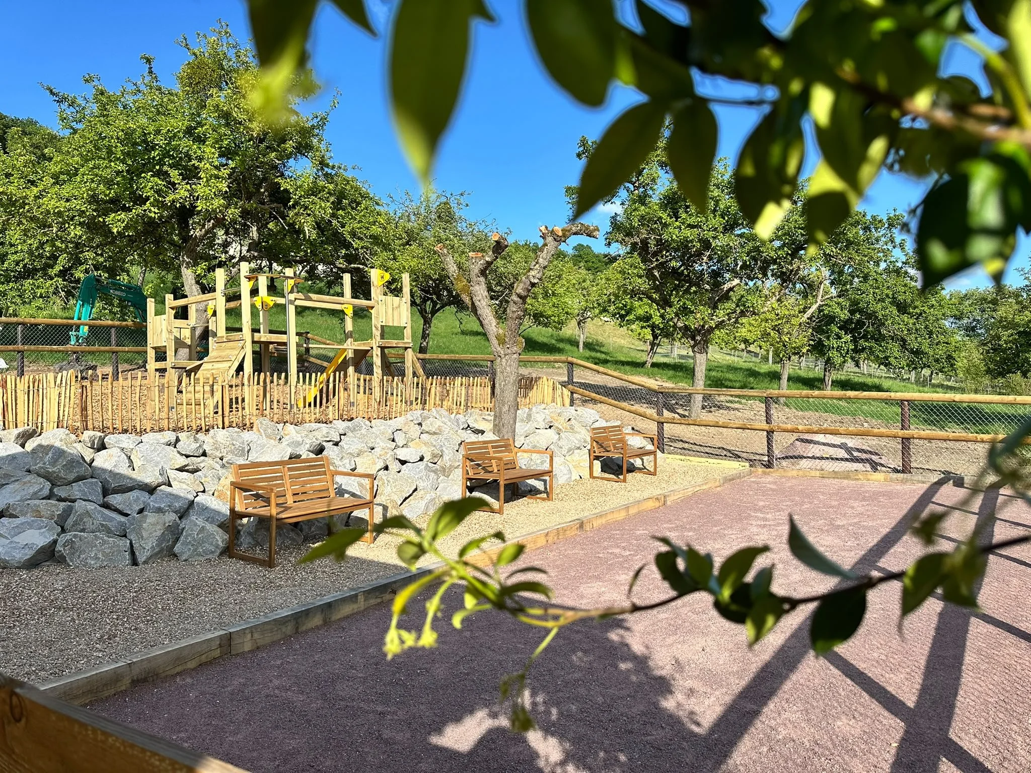 Un parc avec des arbres verts, un espace de jeux en bois, des bancs en bois et une allée en gravier ou terre, sous un ciel bleu ensoleillé.