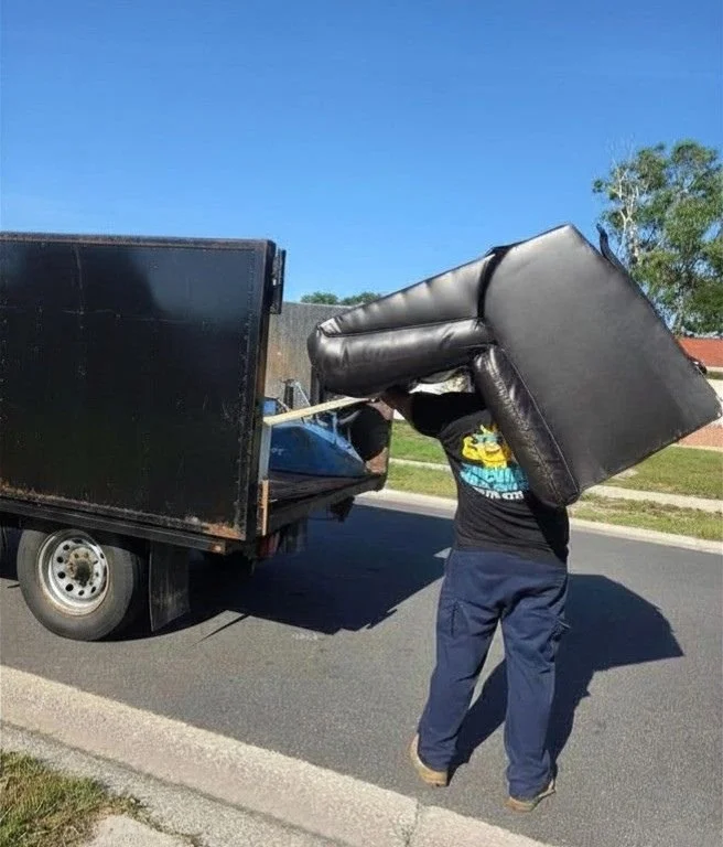 A person carrying a large black furniture piece, possibly a sofa, on their shoulder while walking next to a truck on a residential street.