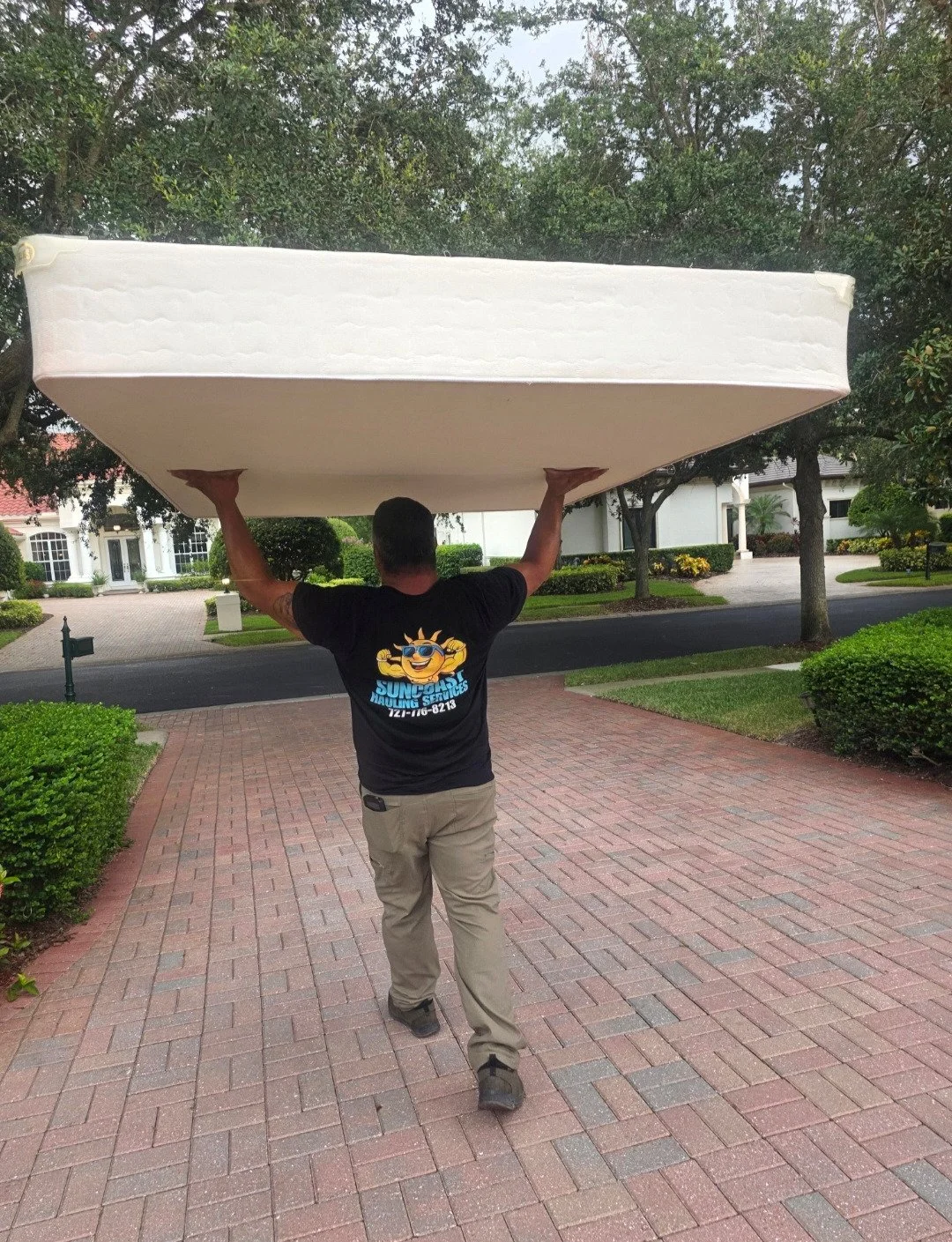 Man carrying a large mattress on his shoulders while walking on a brick pathway in a suburban neighborhood.