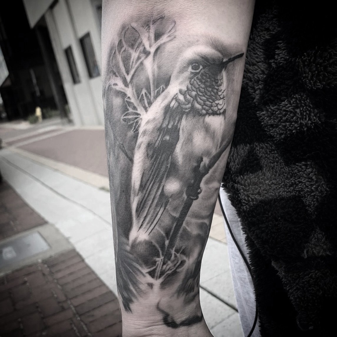 Black and gray tattoo of a hummingbird perched on a branch with leaves on a person's forearm. Tattoo, tattoo artist, tattoos, realistic tattoo, portrait tattoo, artistic tattoo, experienced tattoo artist, Oregon, tattoo artist near me, nature tattoo,