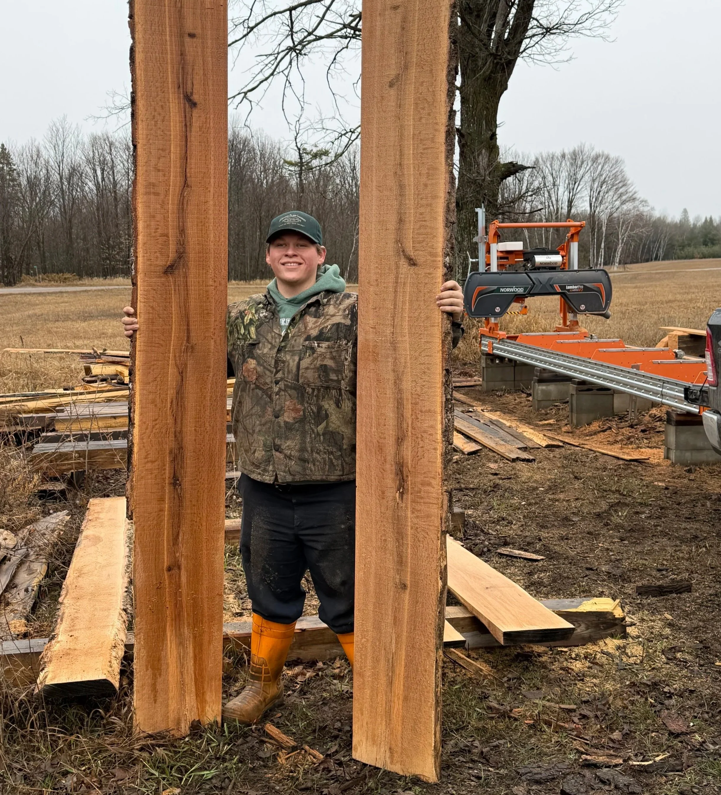 A young man wearing camouflage jacket, green hoodie, and orange rain boots standing behind a large wooden frame outdoors, with a sawmill machine and trees in the background.