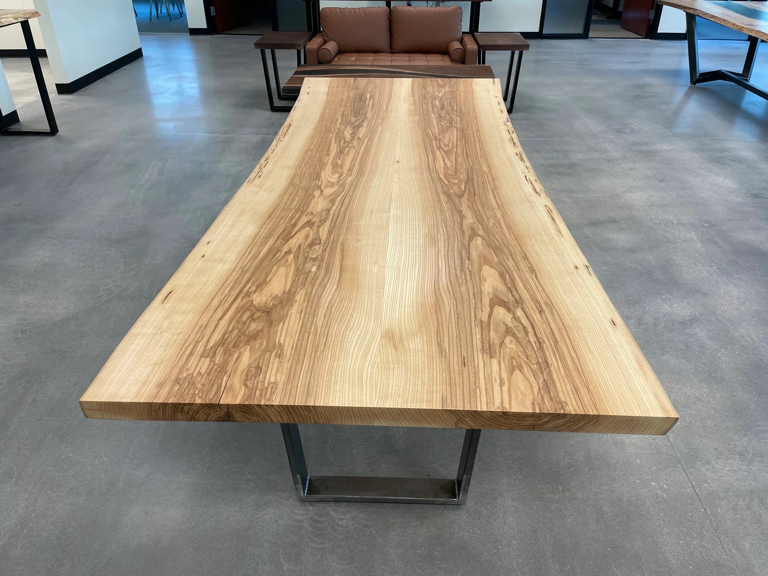 A large wooden table with a natural finish in a modern room, with a brown sofa and additional tables in the background.