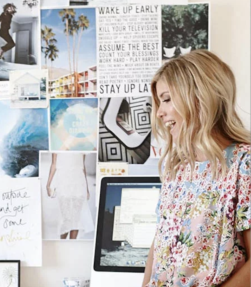 A woman with blonde hair smiling and looking to the right, standing in front of a wall decorated with various photos and a motivational poster, in a room with a computer screen visible.