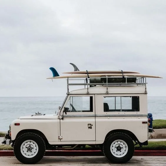 A beige off-road vehicle with surfboards and a kayak on a roof rack parked near the ocean.