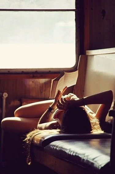 Person lying on a bed reading a book by a window.