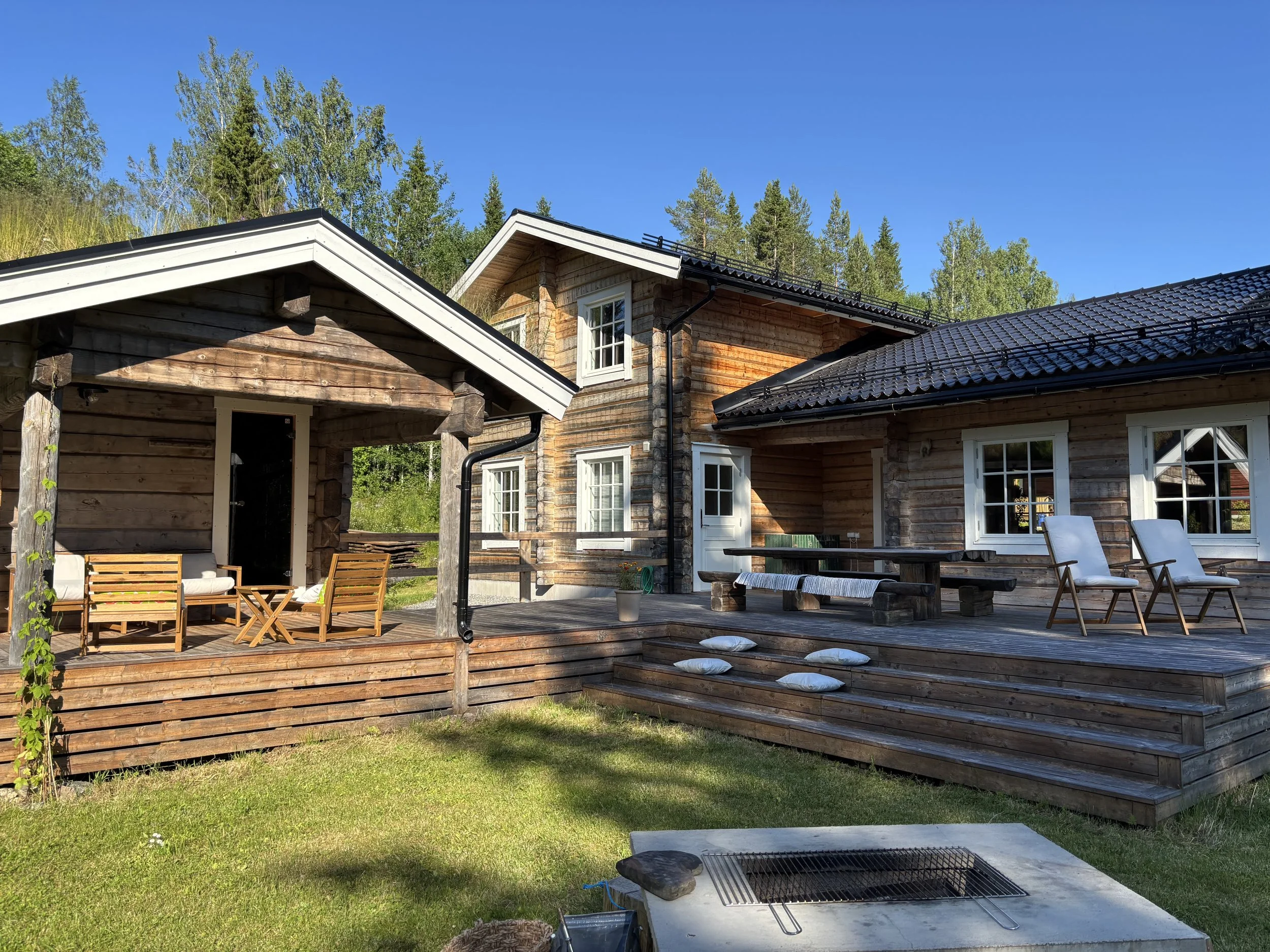 A wooden house with a deck, outdoor seating, and a grassy yard, surrounded by trees under a clear blue sky.
