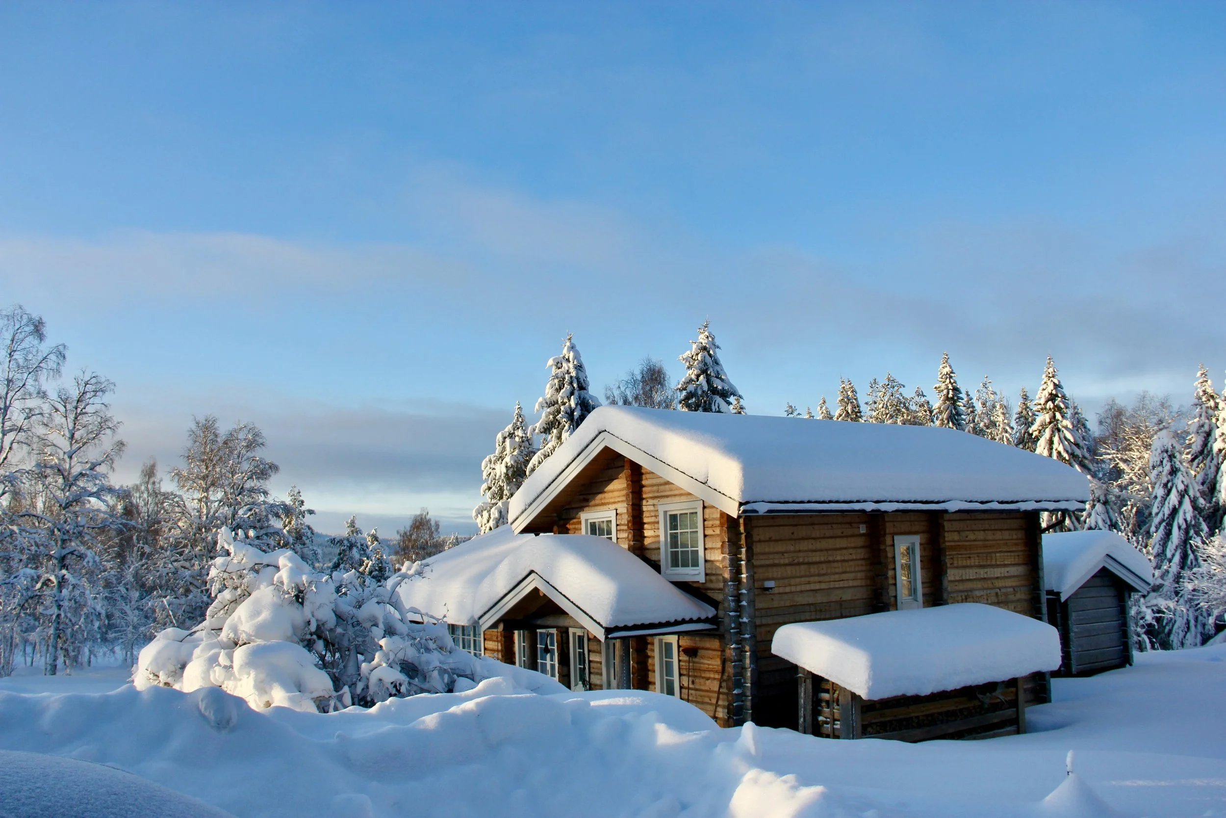 Snow-covered wooden house in a winter landscape with snow-laden trees under a blue sky.