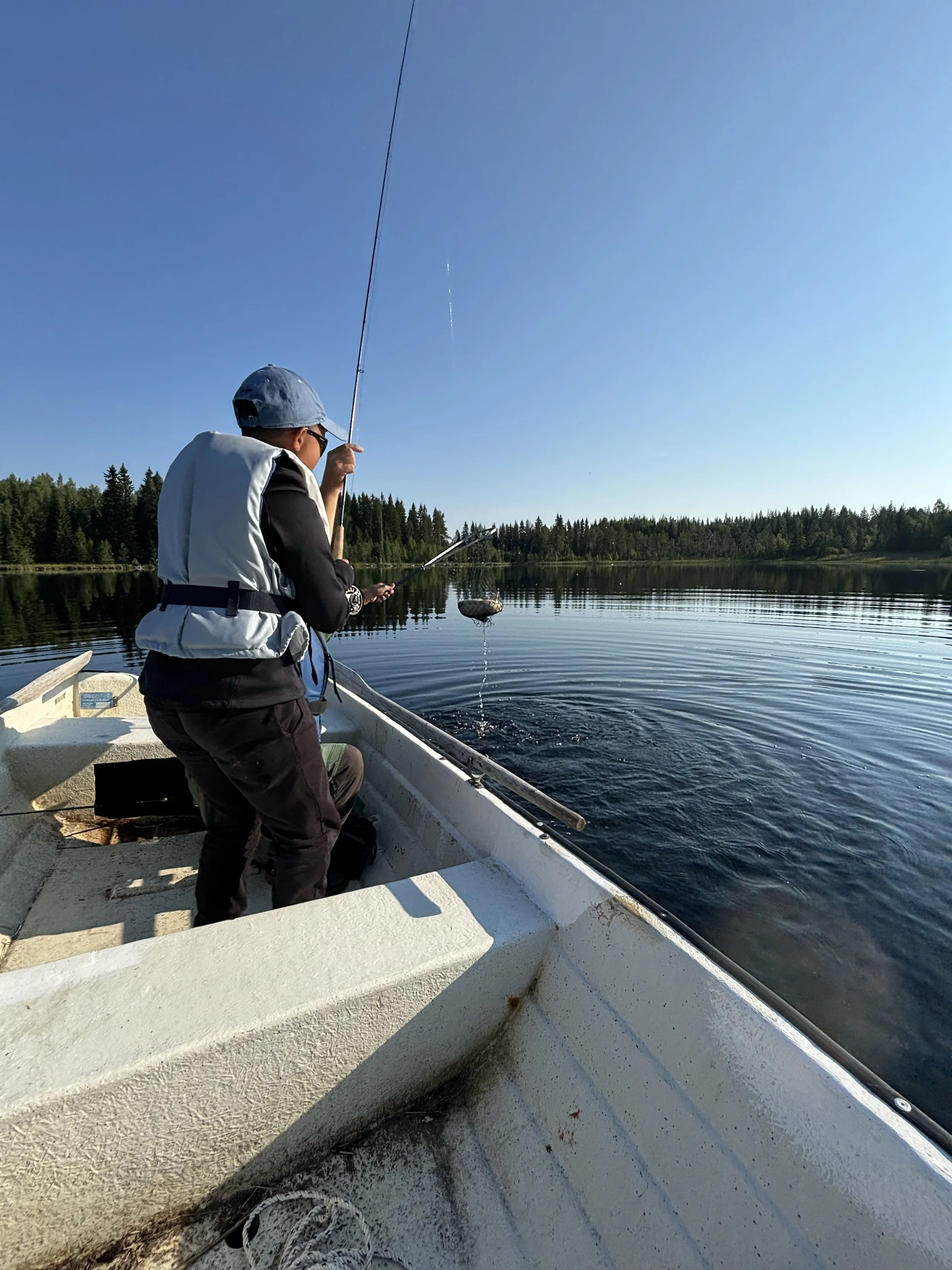 A person fishing from a small boat on a lake with a clear blue sky and forest in the background.