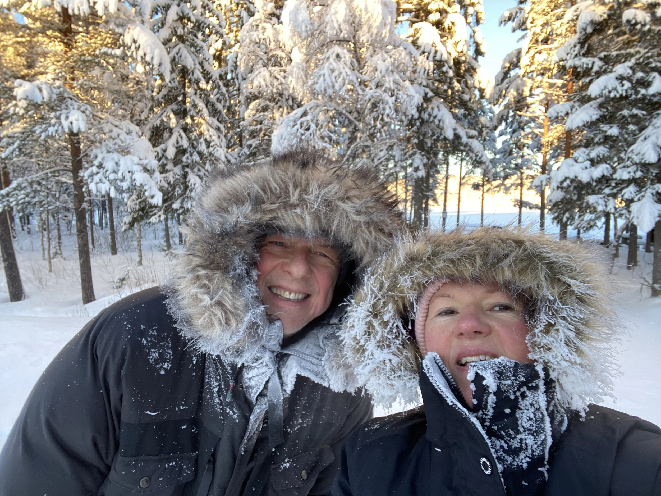 Two people in winter clothing with fur-lined hoods smiling in a snowy forest, with snow-covered trees and a sunset sky in the background.