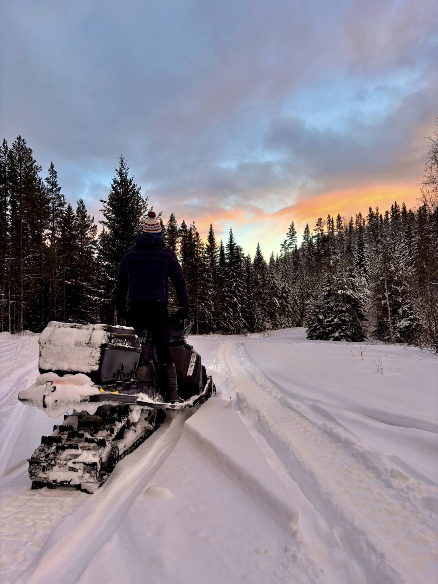 A person riding a snowmobile on a snowy trail in a forest at sunset.