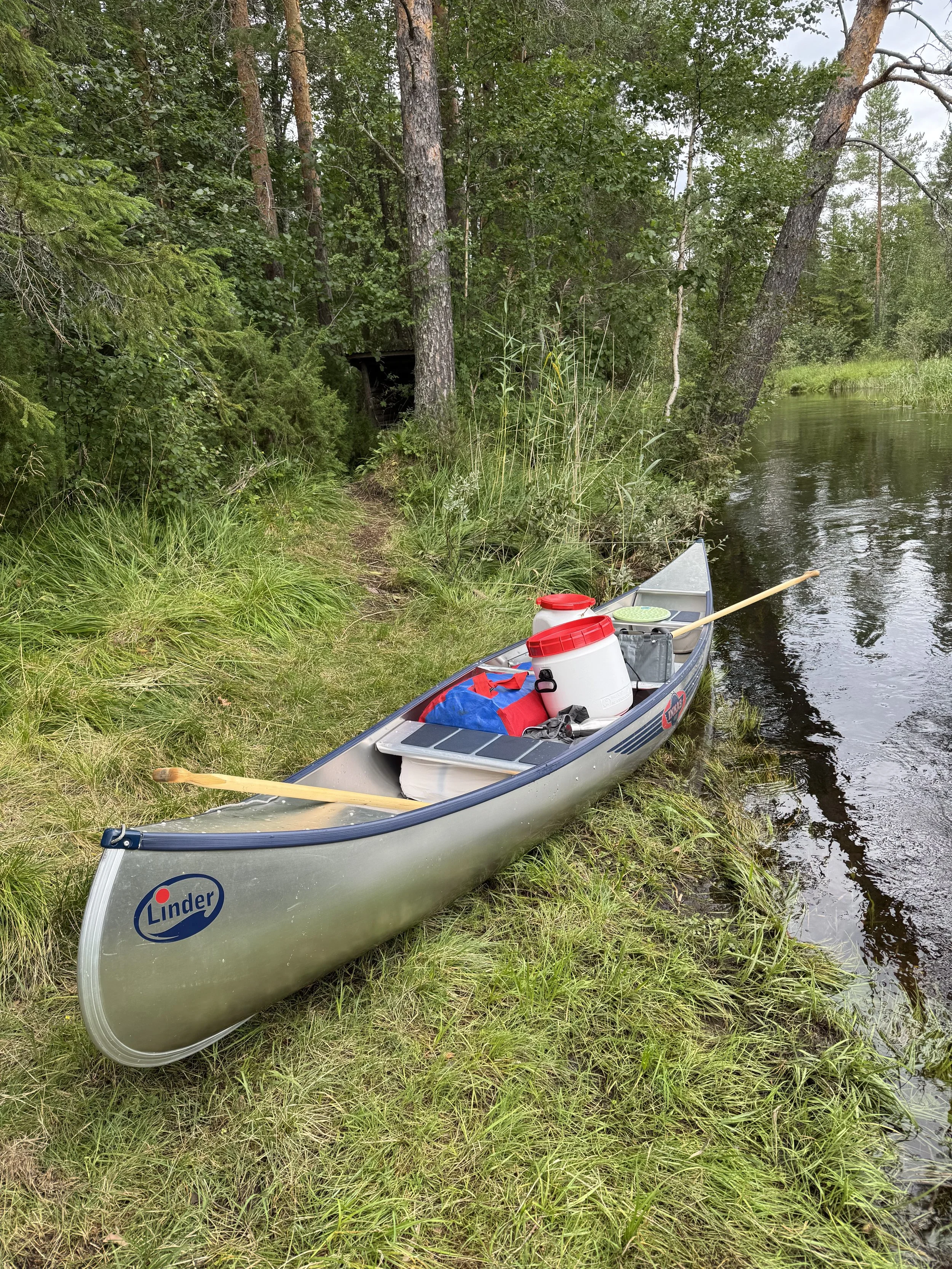 A silver canoe with supplies inside, including plastic water jugs and a paddle, resting on a grassy riverbank surrounded by trees and water.
