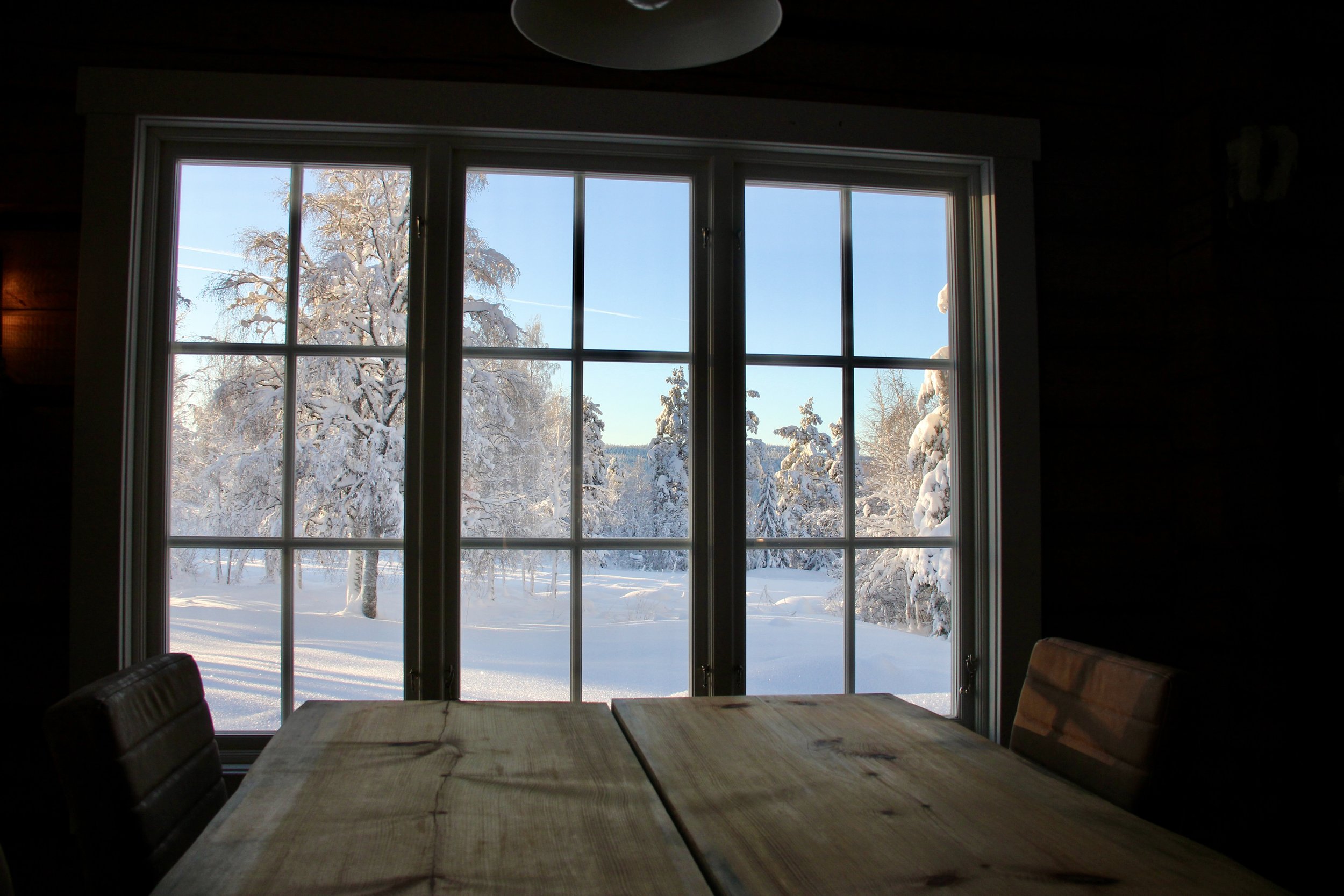 View through a large window showing a winter landscape with snow-covered trees and ground, bright blue sky outside, inside a room with a wooden table and chairs, and dark walls.