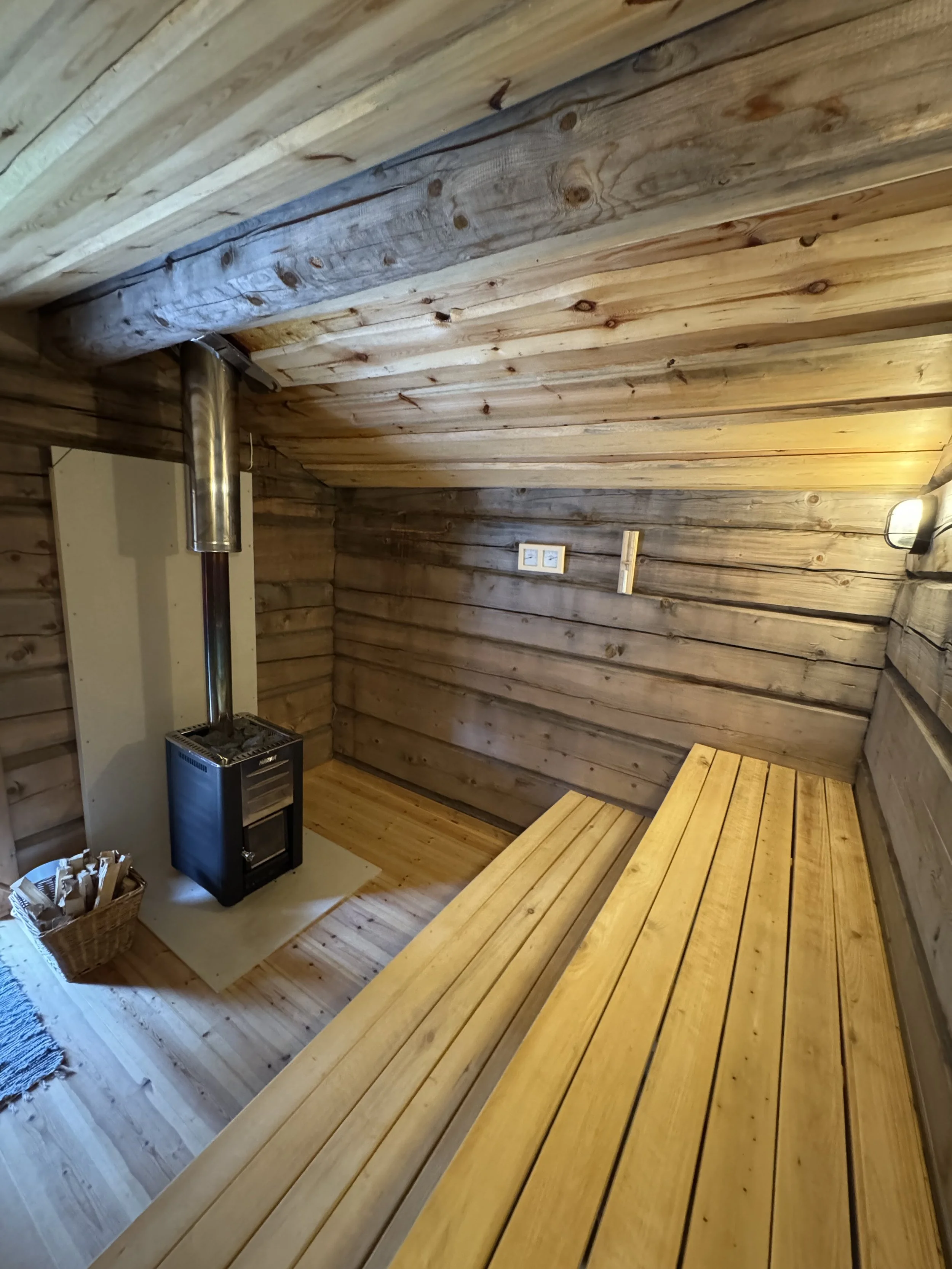 Interior of a small wooden sauna with benches, a wood stove, and wood-paneled walls and ceiling.