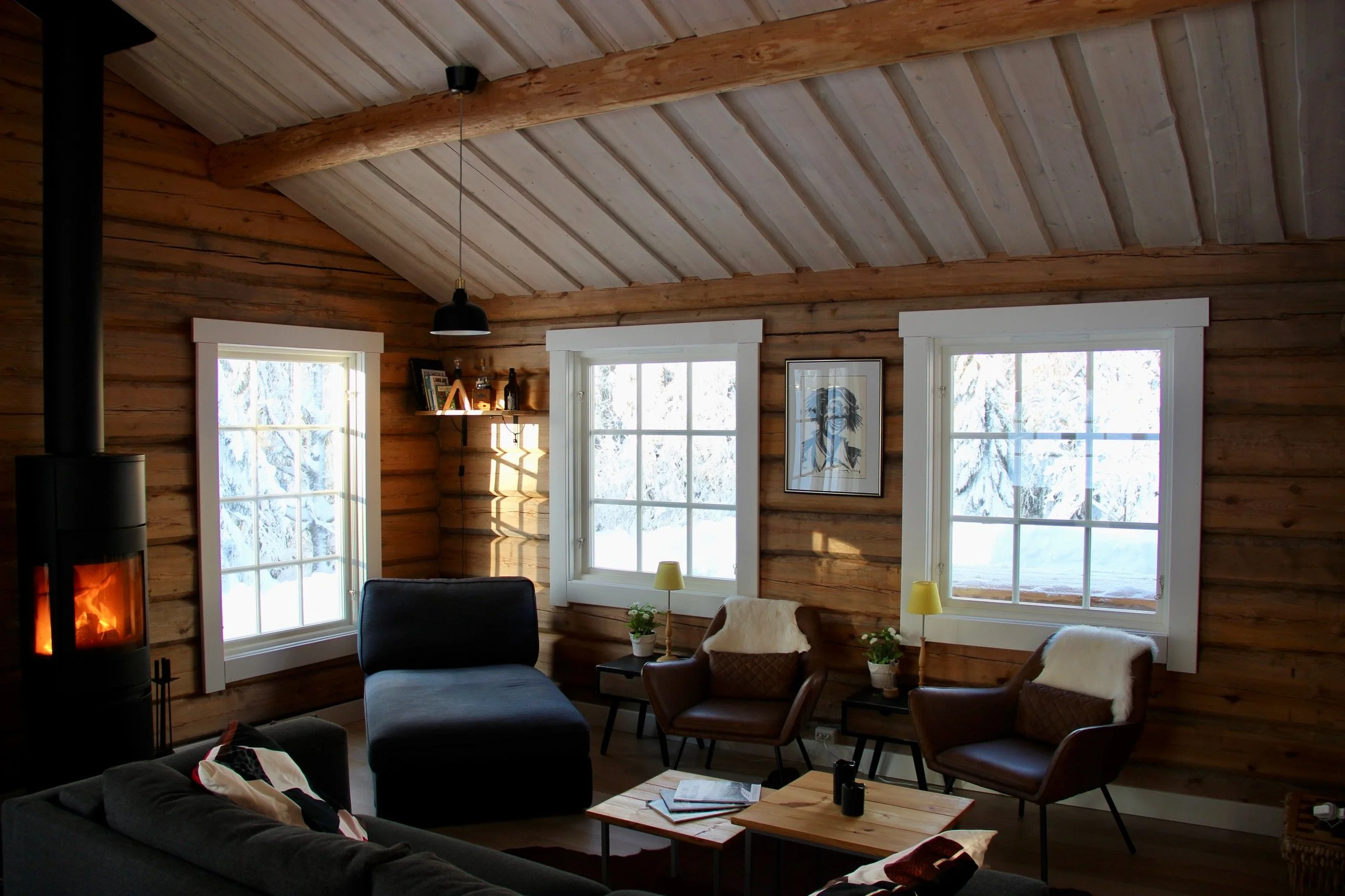 Cozy living room with wooden walls, three windows showing snow outside, black wood stove with visible fire, modern furniture including dark gray sofa, brown leather armchairs with white cushions, small wooden coffee table with books, and side tables 