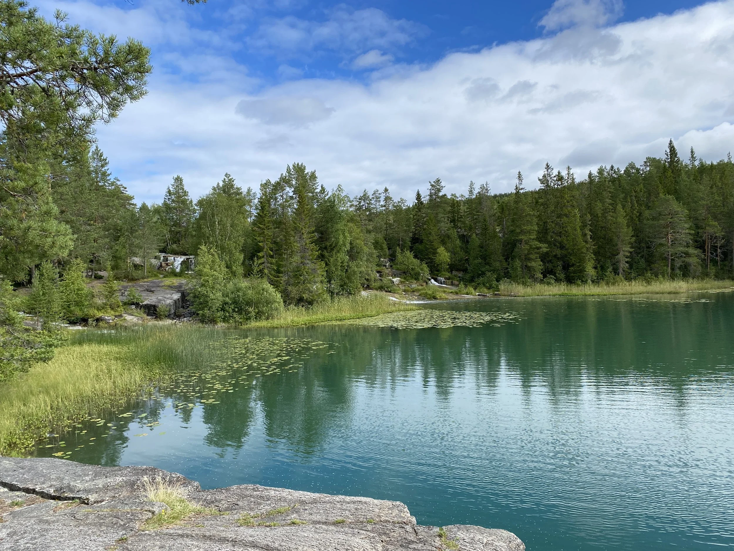 Scenic view of a lake surrounded by lush green trees and rocks, with a partly cloudy sky above.