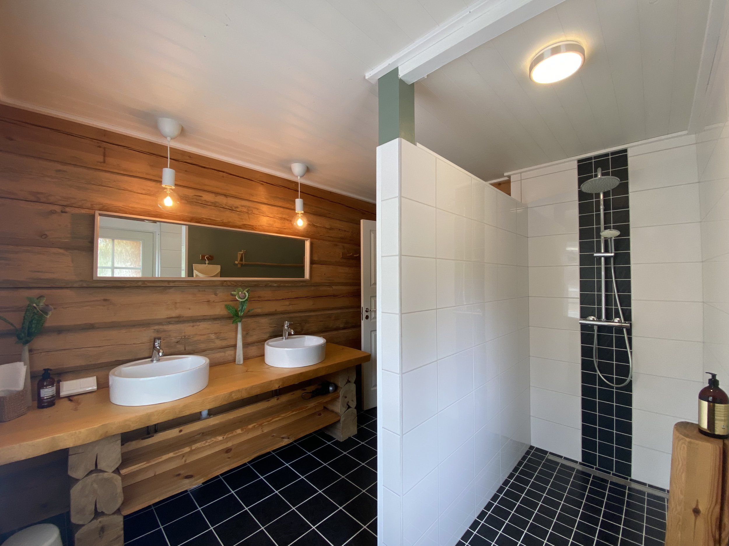 A modern bathroom with a double wooden vanity with two oval sinks, decorative plants, a large mirror, and a shower area with white tiles and a black accent strip, all under warm lighting.