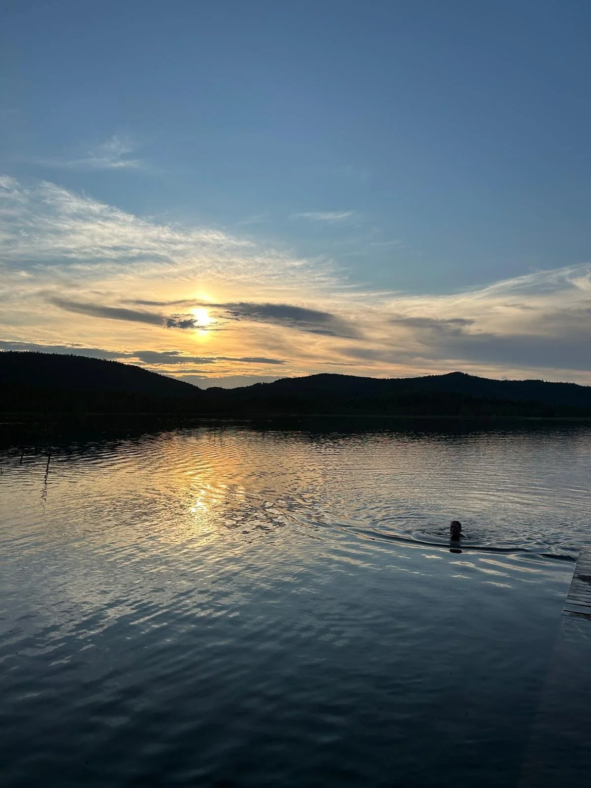 A person swimming in a calm lake during sunset with a scenic mountain range in the background and a partly cloudy sky.