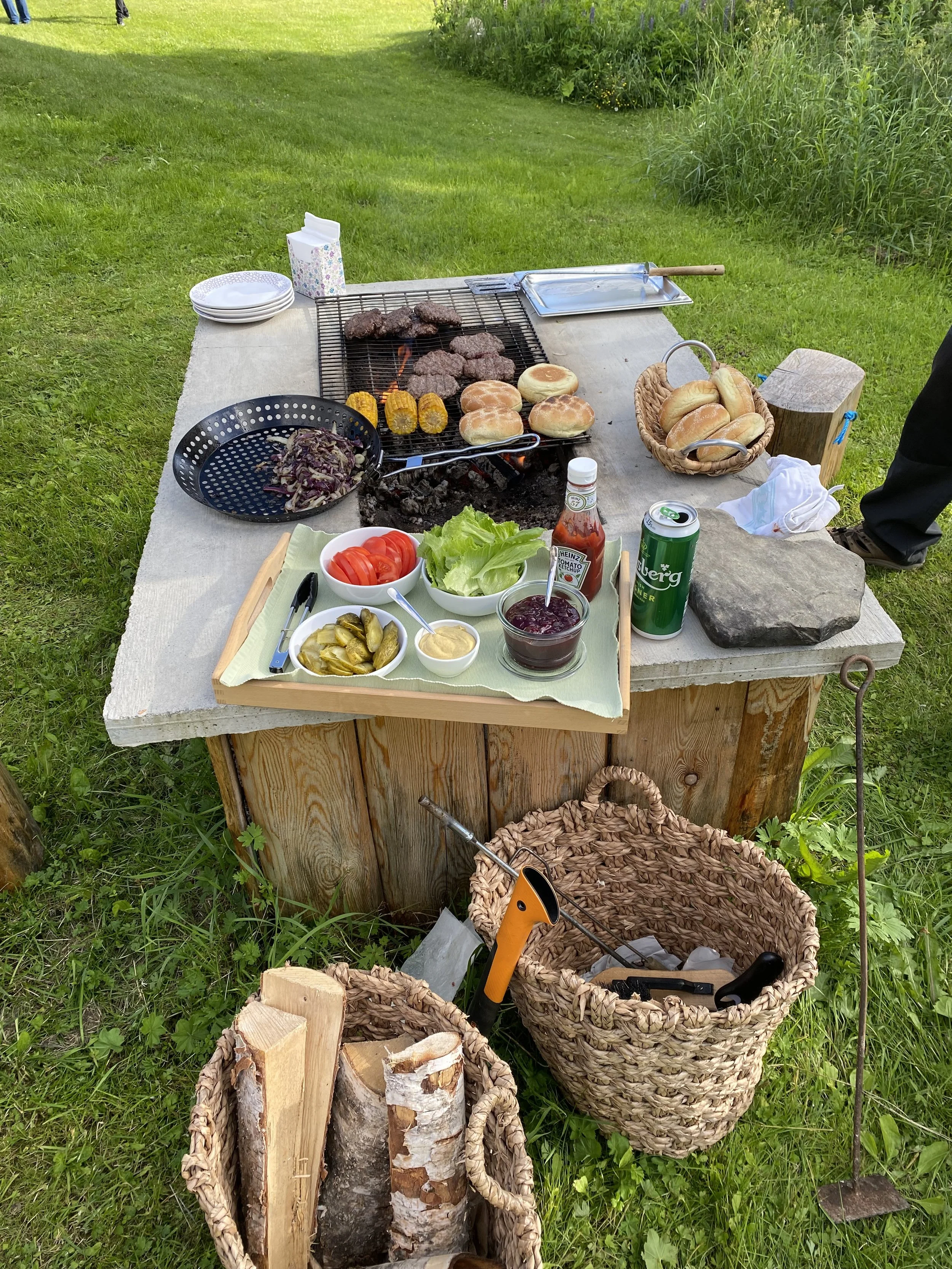 Outdoor barbecue setup with grilling meats, hamburger buns, and vegetables, along with side dishes including sliced tomatoes, pickles, and berry jam, all arranged on a wooden table. There are baskets of bread, a container of firewood, and grilling tools, with a grassy yard and plants in the background.