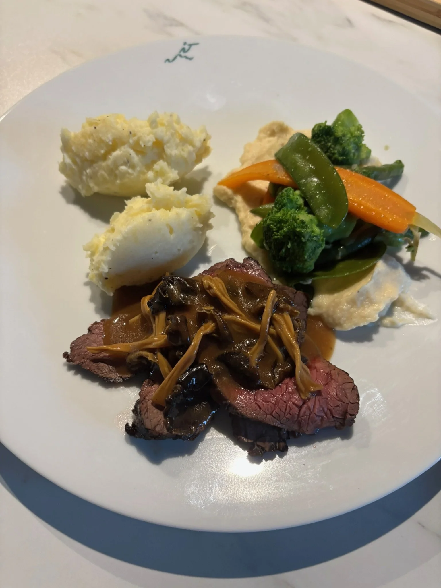 A plate with slices of beef topped with mushroom gravy, mashed potatoes, and a serving of mixed vegetables including broccoli, carrots, and green beans.