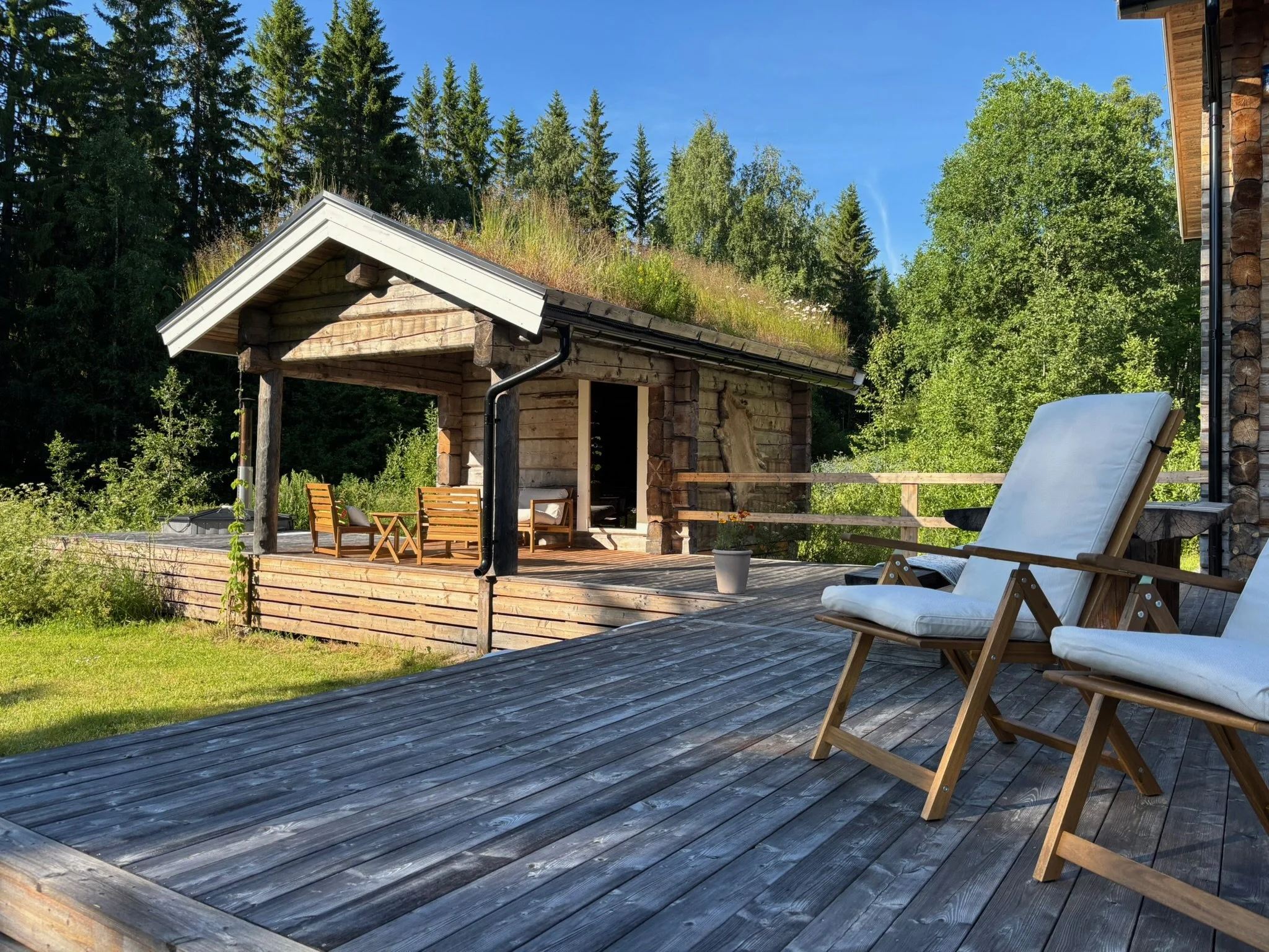 A wooden deck overlooking a lush green forest with a small wooden cabin in the background. The deck has white cushioned chairs and a small pot with a plant.