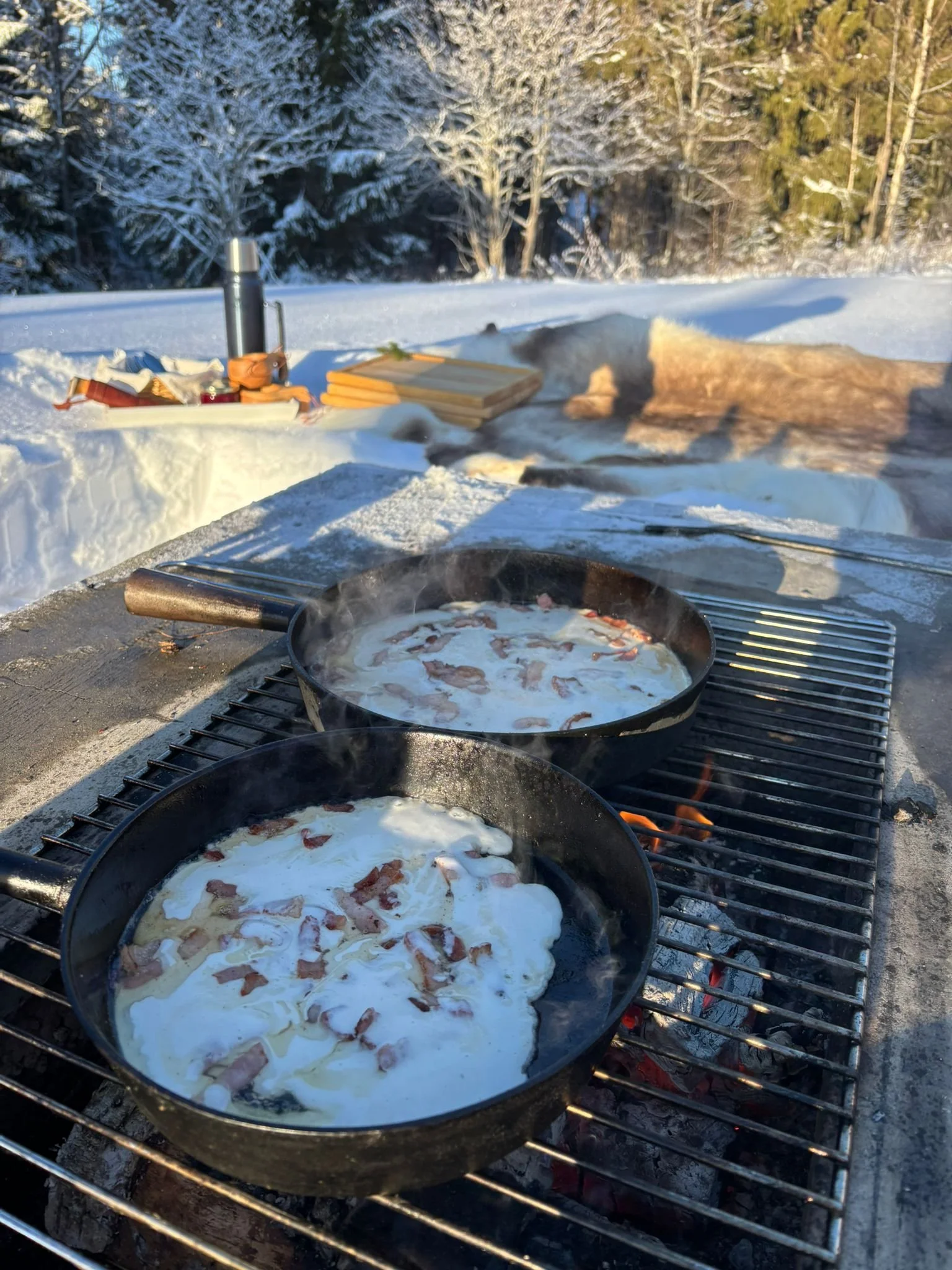 Two frying pans on an outdoor grill in a snowy landscape, cooking pancakes with bacon, with snow-covered trees and a table with supplies in the background.
