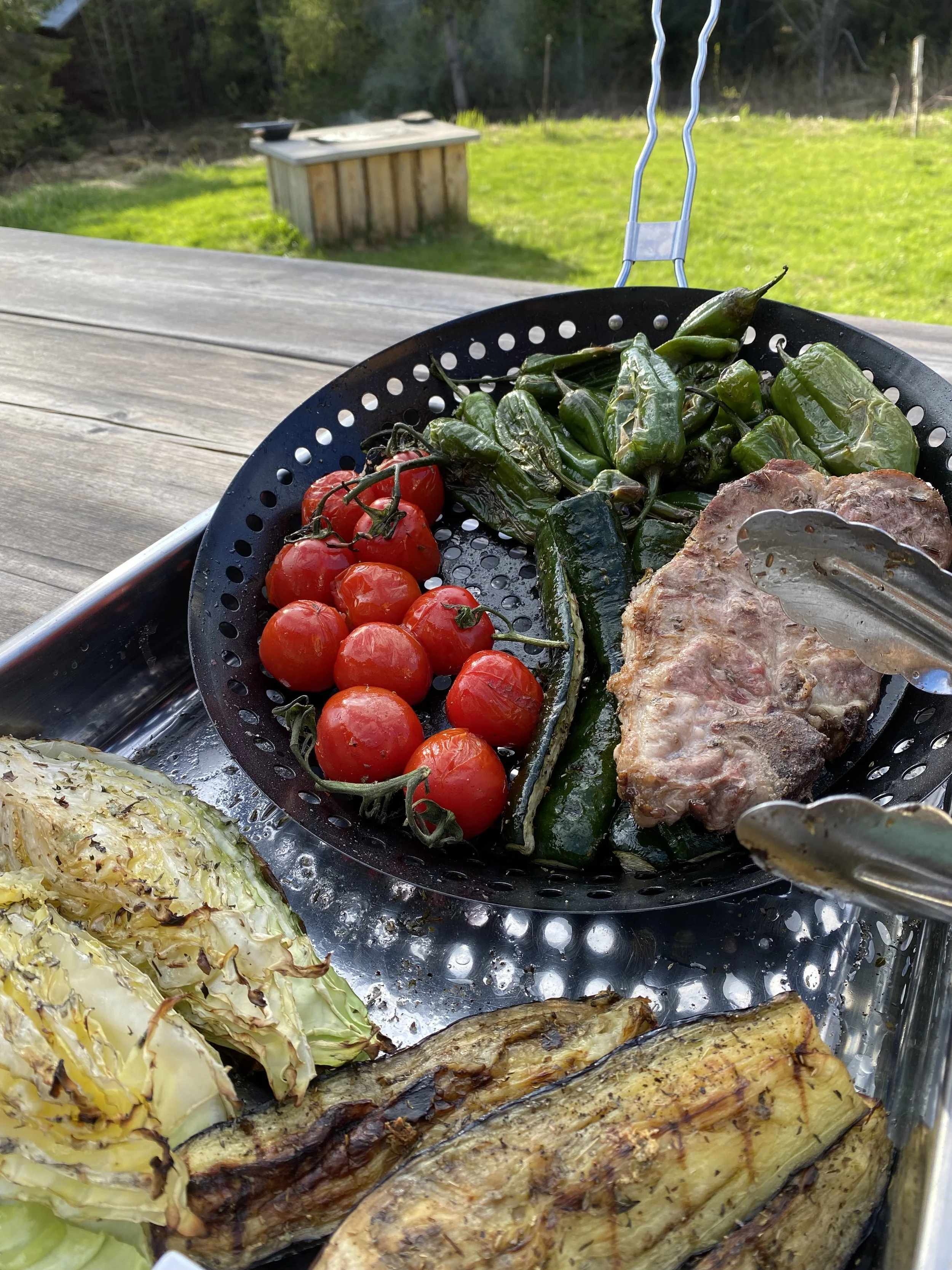 Fresh cherry tomatoes, green peppers, and a cooked steak in a black colander on a wooden table outdoors.