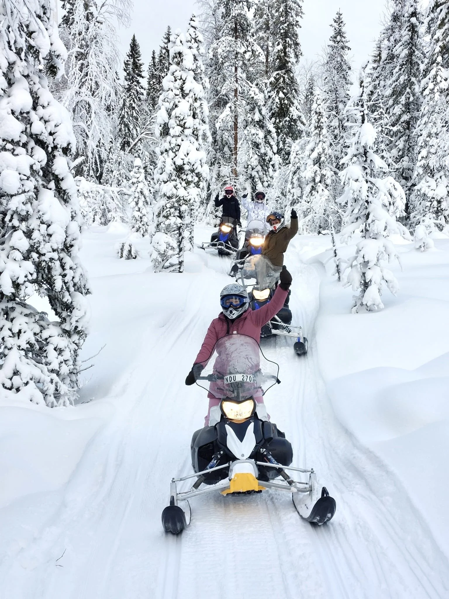 A group of people riding snowmobiles through a snowy forest with snow-covered trees. The person in front is waving, wearing a pink jacket and snow goggles. Others follow behind, all dressed in winter gear.