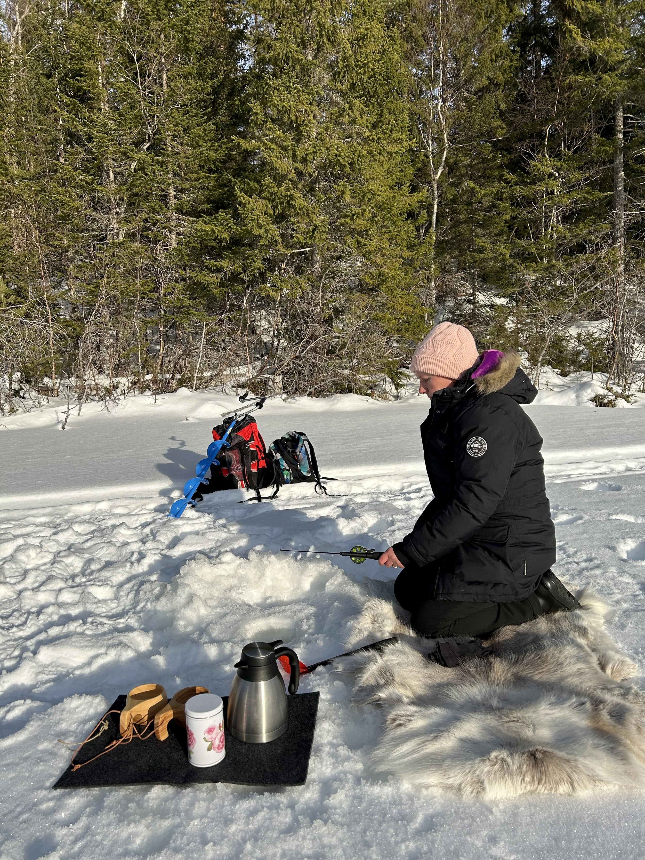 A woman in a black winter jacket kneeling on snow in a forest, fishing through a hole in the ice, with a fur blanket, thermos, mug, and sunglasses nearby and an ice auger and backpack behind her.