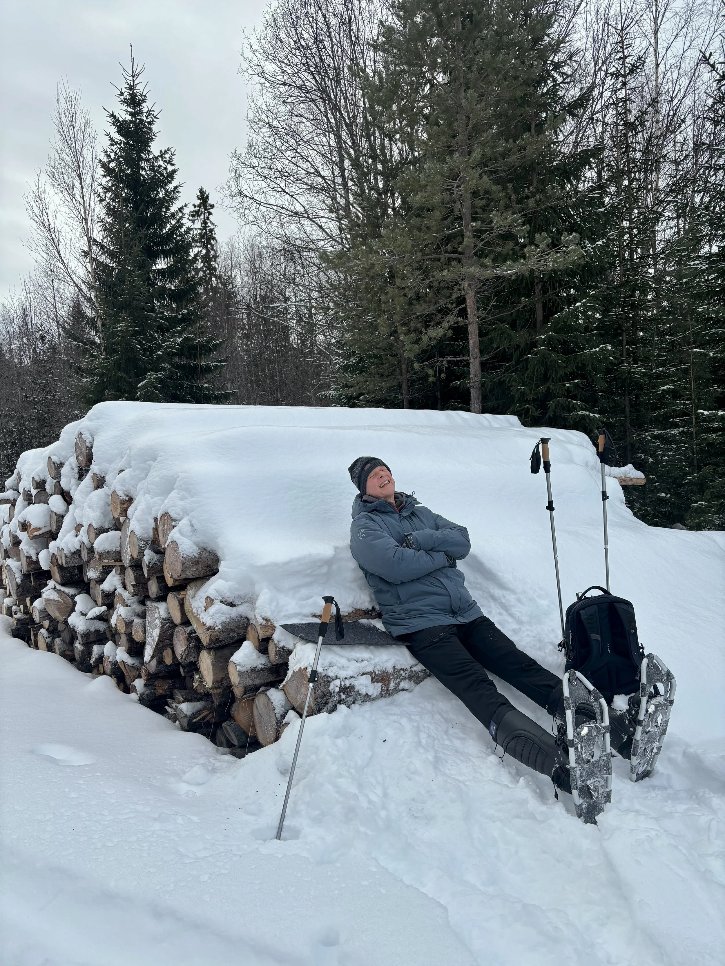 A person wearing a winter jacket and hat relaxing on a snow-covered log in a snowy forest, with snowshoes and trekking poles nearby.