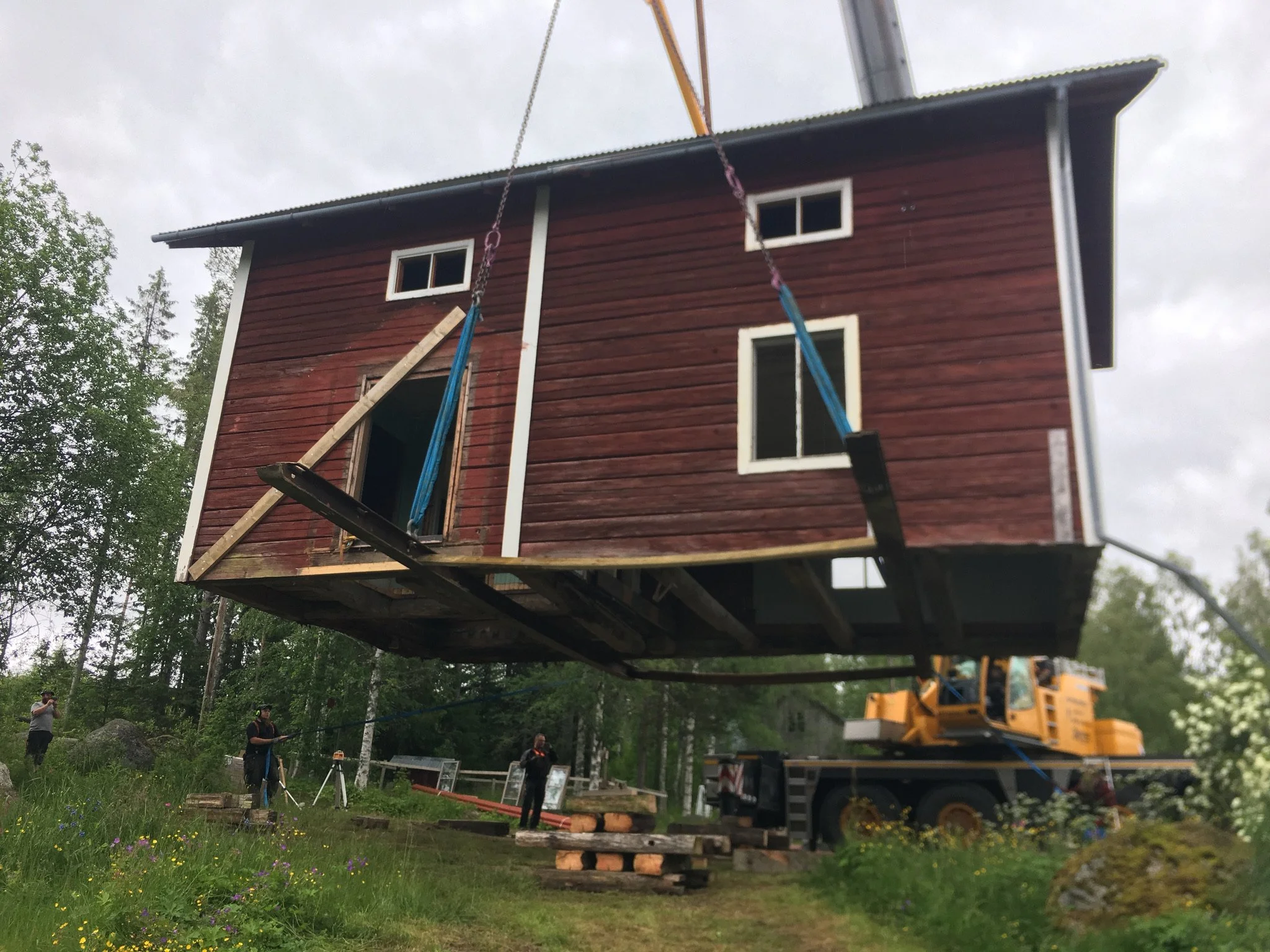 A large yellow crane lifting a small wooden house in a forested area with people observing.