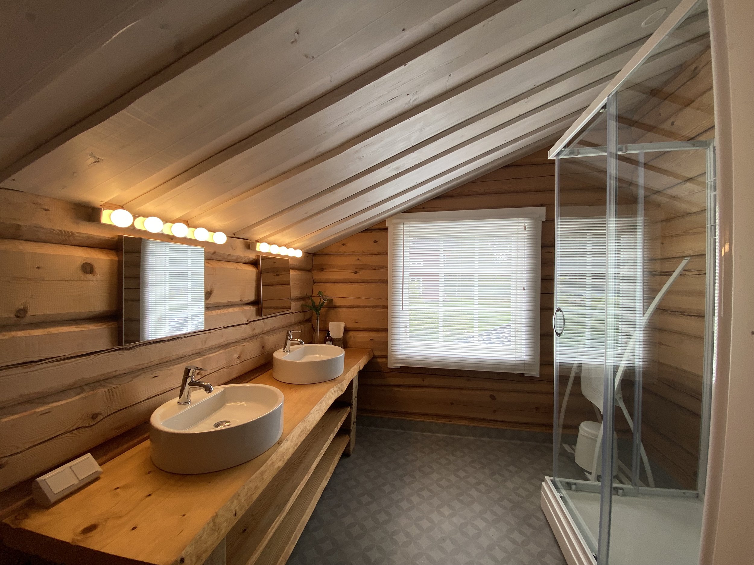 A rustic bathroom with wooden walls and ceiling, featuring two white vessel sinks on a wooden countertop, a window with blinds, and a glass-enclosed shower with a showerhead.