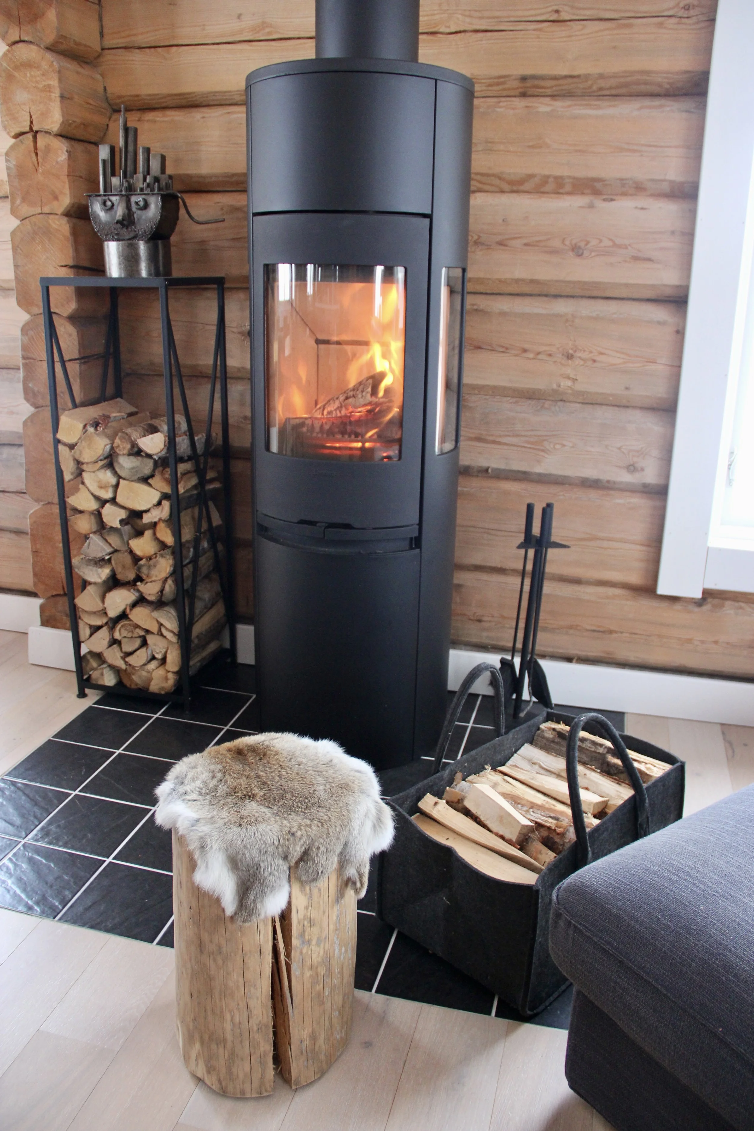 Modern wood-burning stove with a glass front showing a fire, surrounded by firewood and cozy decor in a log cabin interior.