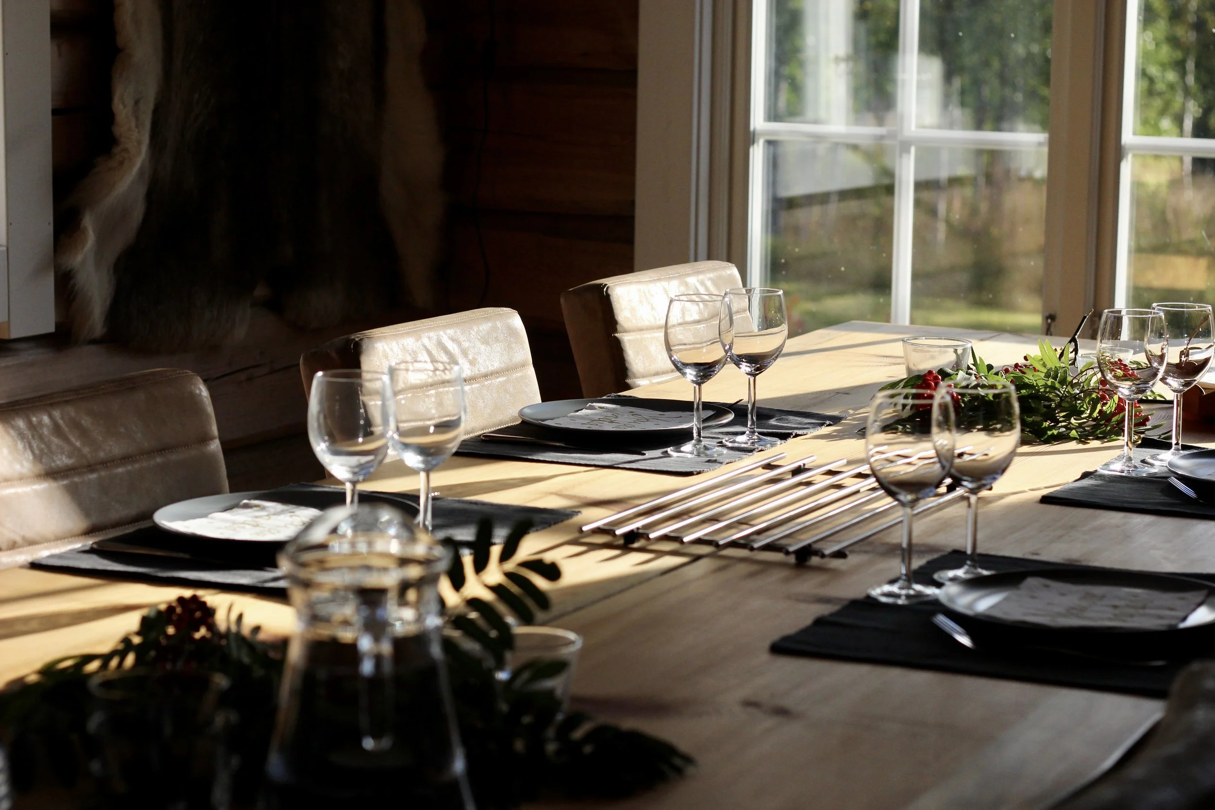 A dining table set with plates, wine glasses, and cutlery, decorated with greenery and placed near large windows letting in natural sunlight.