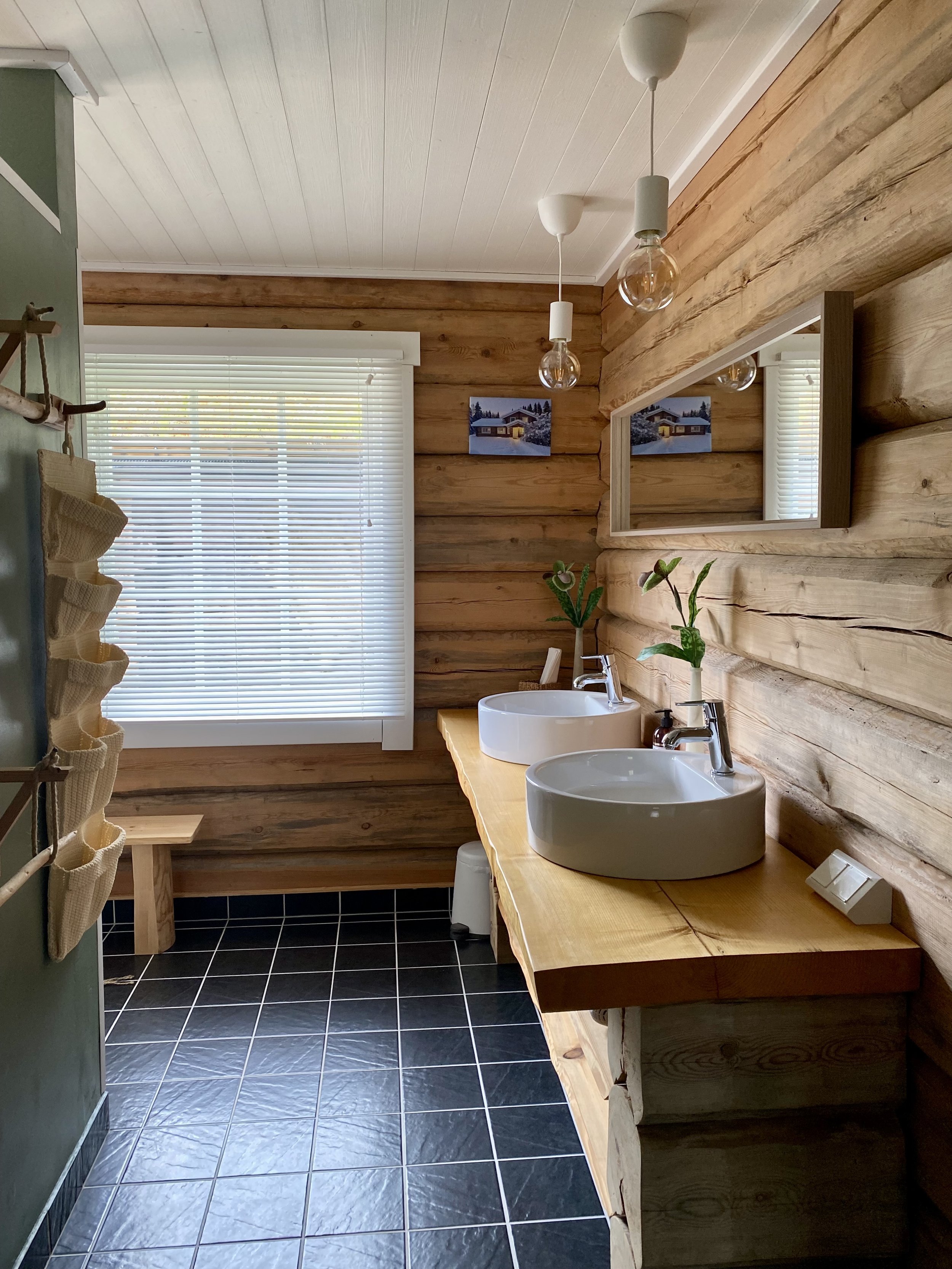 Bathroom with wooden walls, black tiled floor, and two white vessel sinks on a natural wood countertop. There are small plants in vases and a mirror above the sinks. A window with blinds lets in natural light. Modern light fixtures hang from the ceil