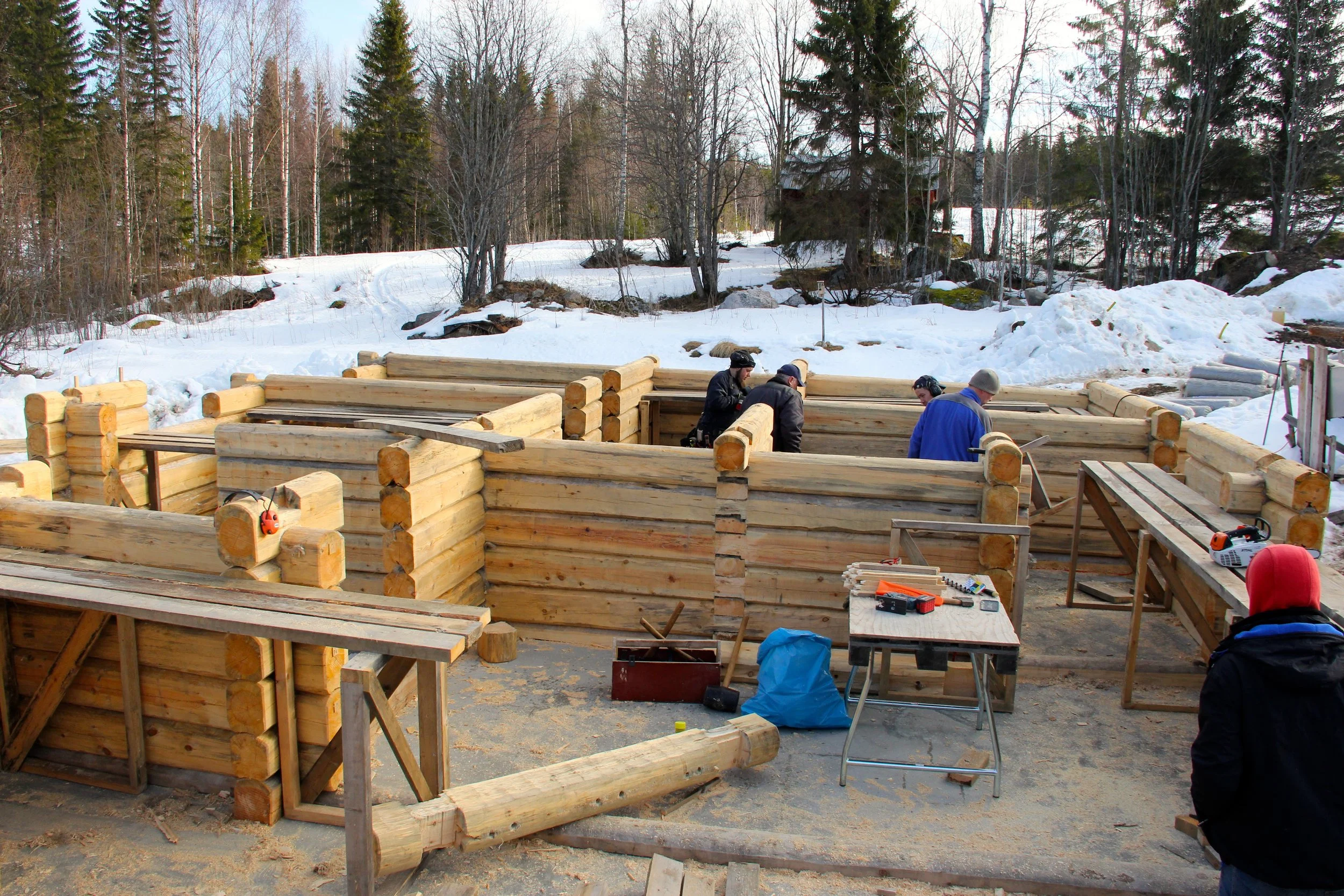 Workers constructing a log cabin with snow on the ground and trees in the background.