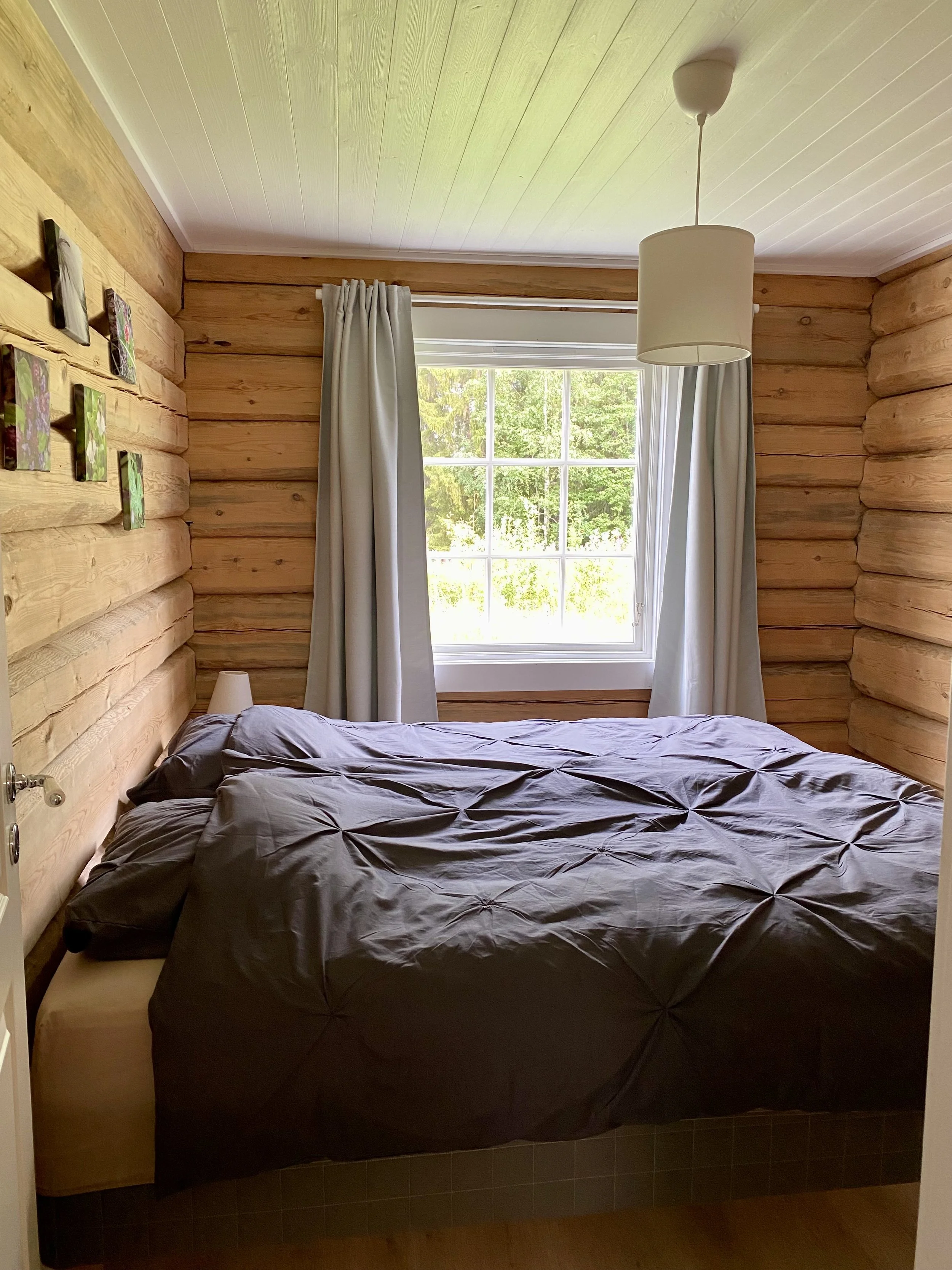 A cozy bedroom with log cabin style wooden walls, a large window with white curtains, and a bed with a dark gray comforter and matching pillows. A small bedside lamp is beside the bed, and a simple ceiling light fixture hangs overhead.