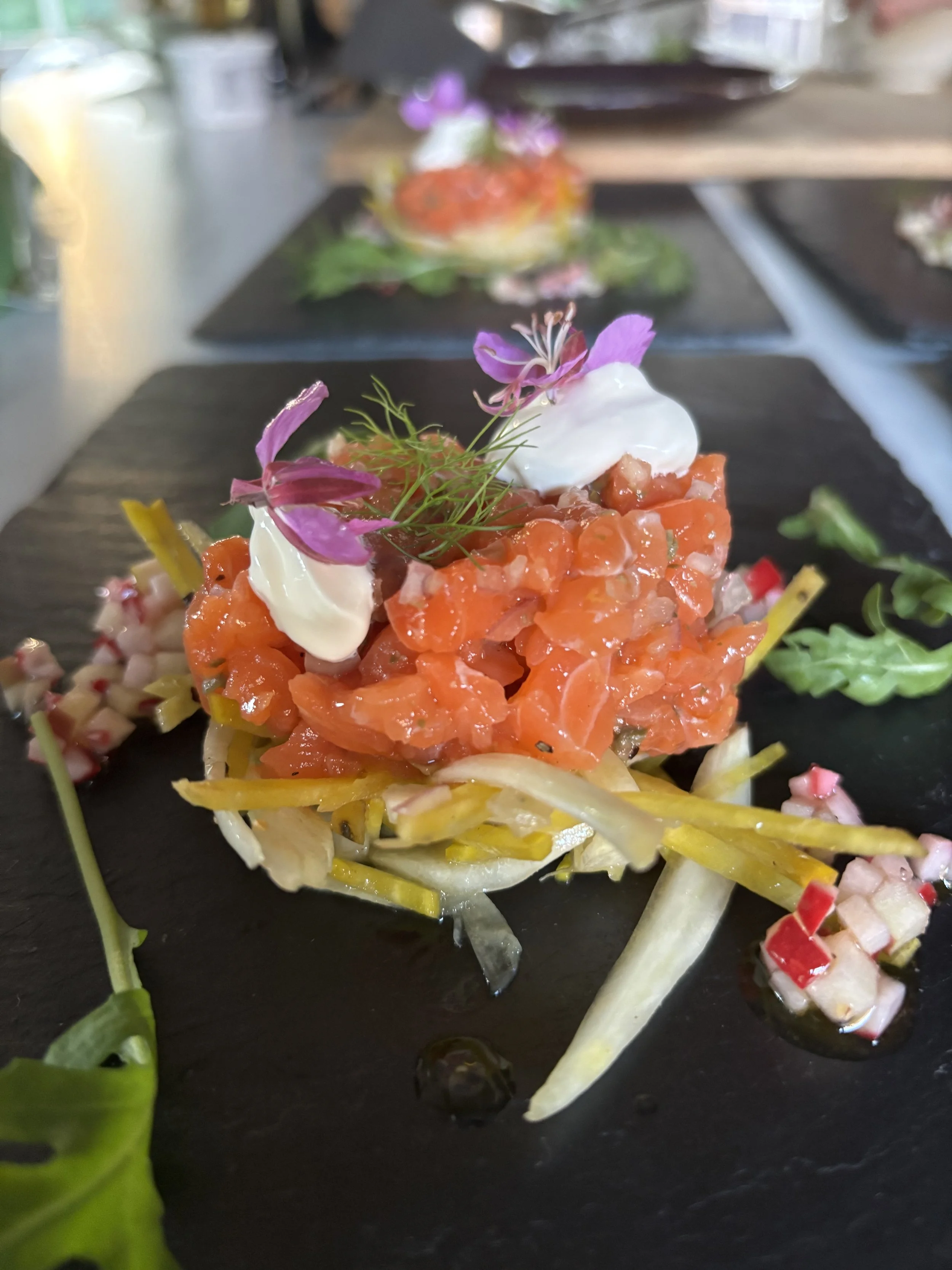 Close-up of a plated dish with diced raw salmon, garnished with microgreens and edible flowers, served on a bed of julienned vegetables on a black slate plate.