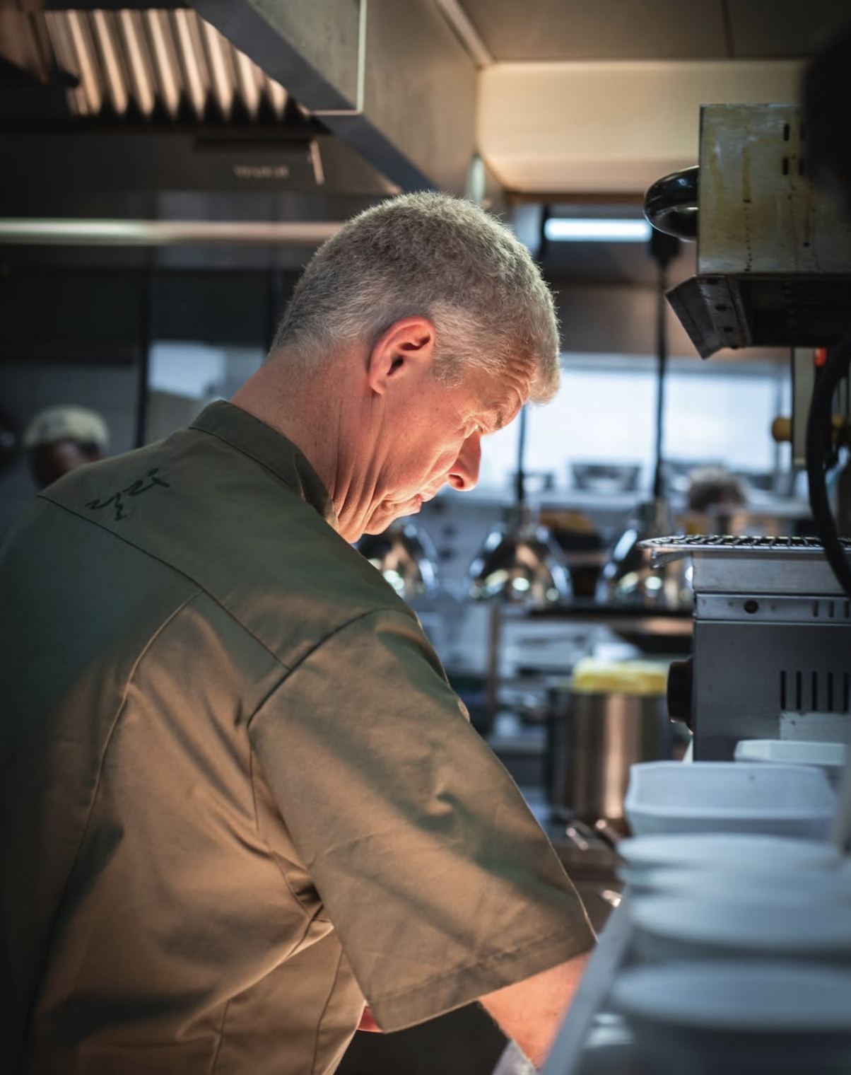 A chef in a commercial kitchen preparing food, with kitchen equipment and shelves in the background.