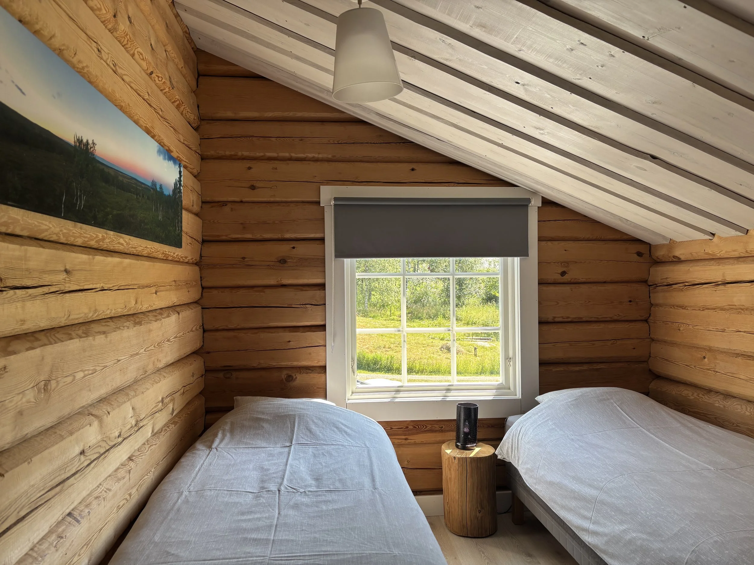 A cozy bedroom with wooden log walls, featuring two beds with white bedding, a window with a gray roller blind overlooking a green outdoor landscape, a round wooden side table between the beds, and a landscape painting on the left wall.