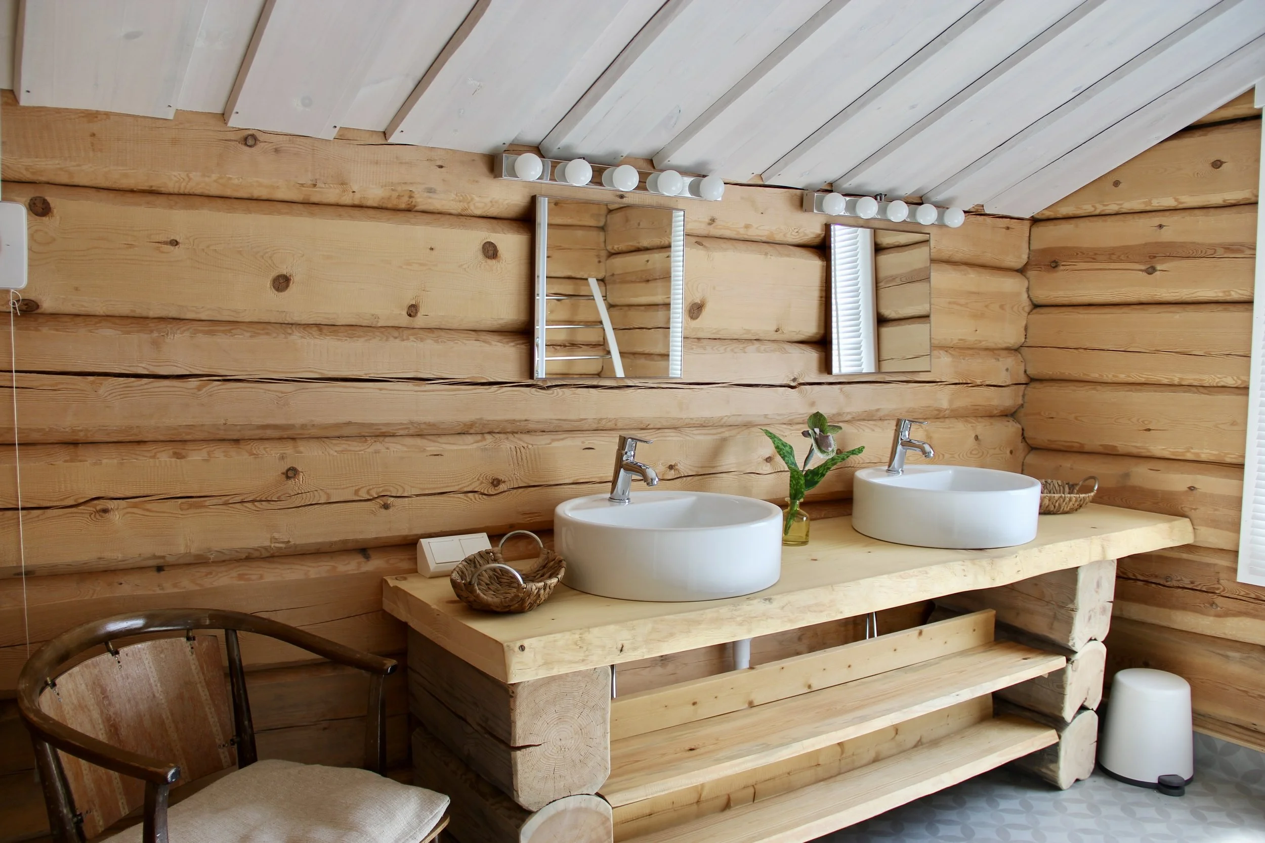 Wooden bathroom vanity with two white vessel sinks, a potted plant, baskets, and mirrors, set against log cabin walls and ceiling.