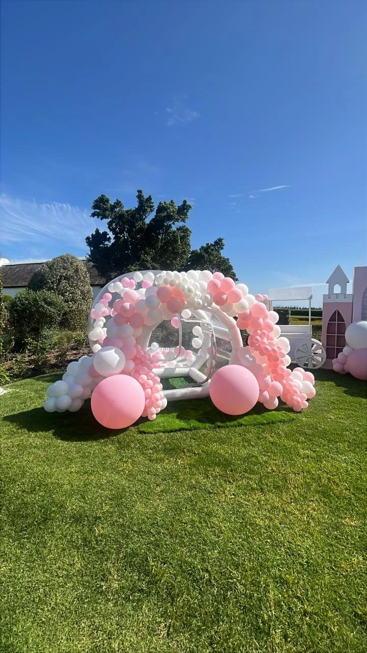 A decorated outdoor setting featuring a white balloon carriage adorned with pink and white balloons, set on green grass under a clear blue sky.
