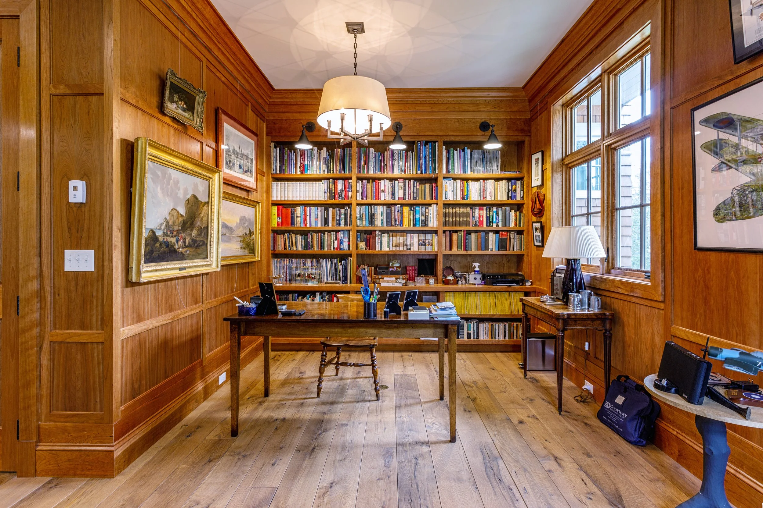 Wood-paneled study with bookshelves, paintings, and a wooden desk.