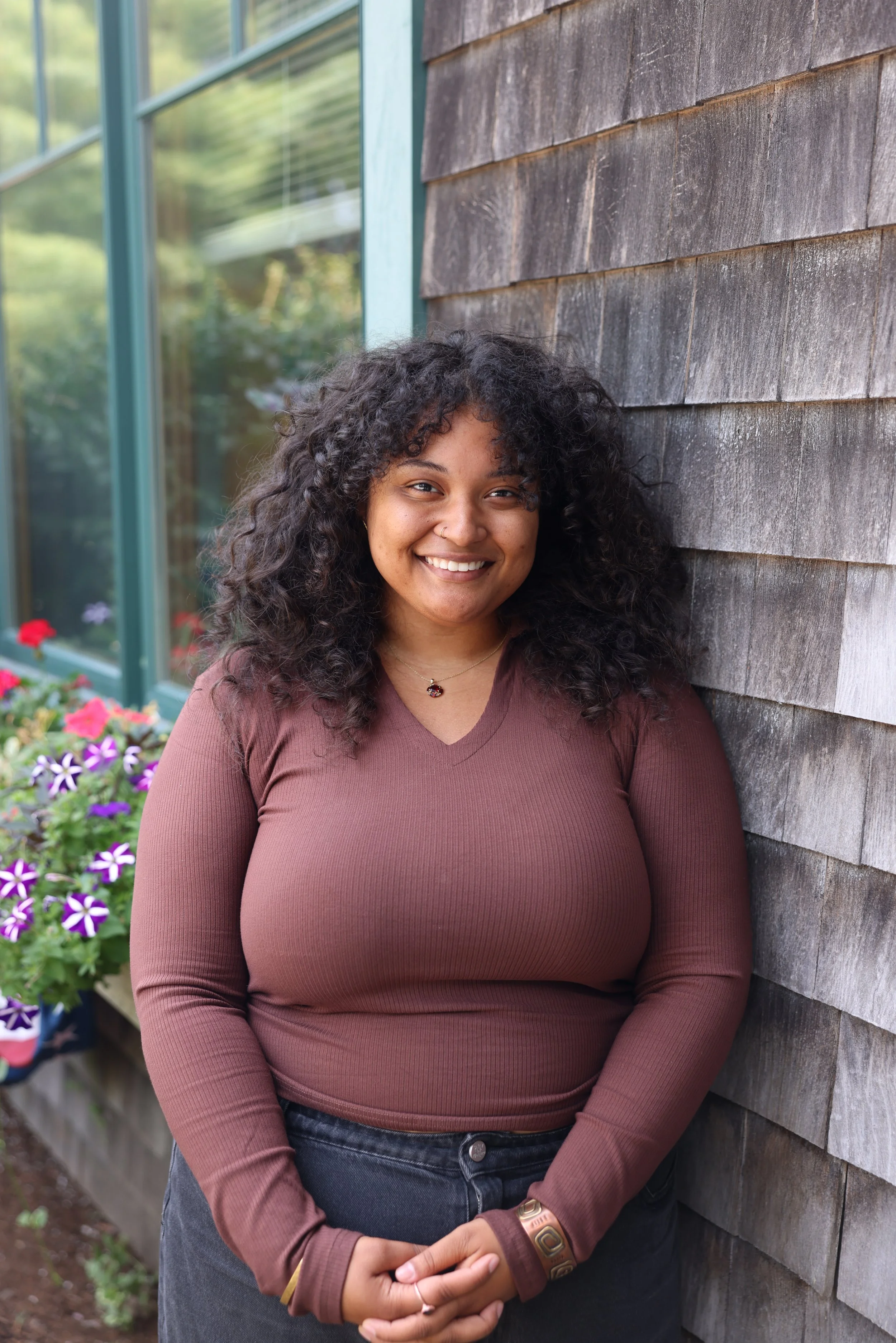 Person with curly hair and glasses smiling while sitting on a wooden bench indoors, wearing a black top and beige pants.