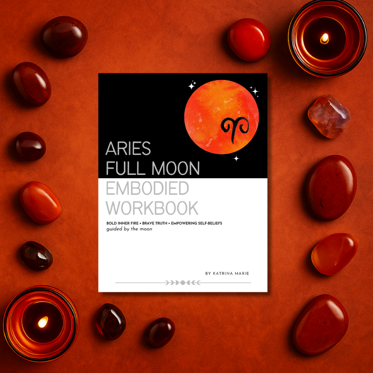 The Aries Full Moon Embodied Workbook cover displayed on a burnt orange surface surrounded by red jasper, carnelian, garnet, and lit candles — bold inner fire, brave truth, empowering self-beliefs, guided by the moon, by Katrina Marie