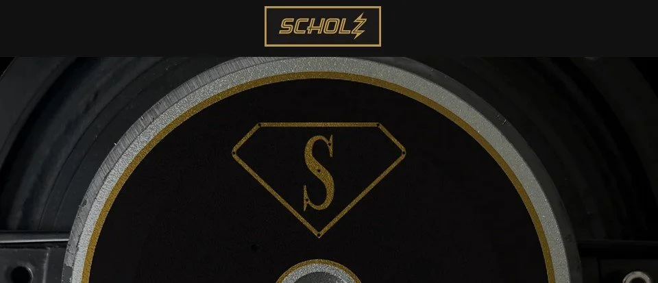 Close-up of a black and gold weight plate with a diamond-shaped emblem and the letter 'S' in the center, branded 'Scholz' at the top.