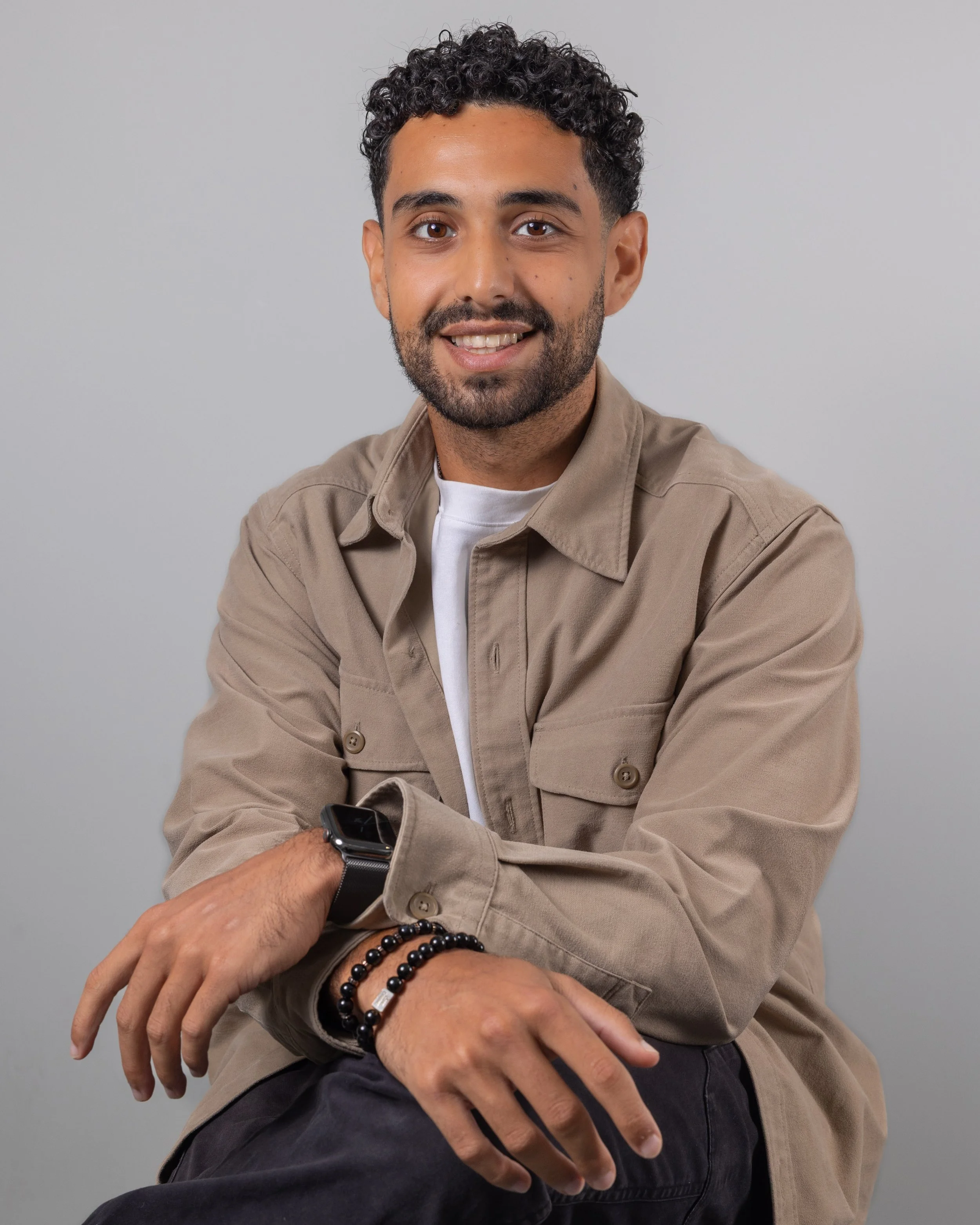 A young man with curly dark hair and a beard smiling, wearing a tan jacket and a white T-shirt, sitting with his arms crossed against a plain gray backdrop.