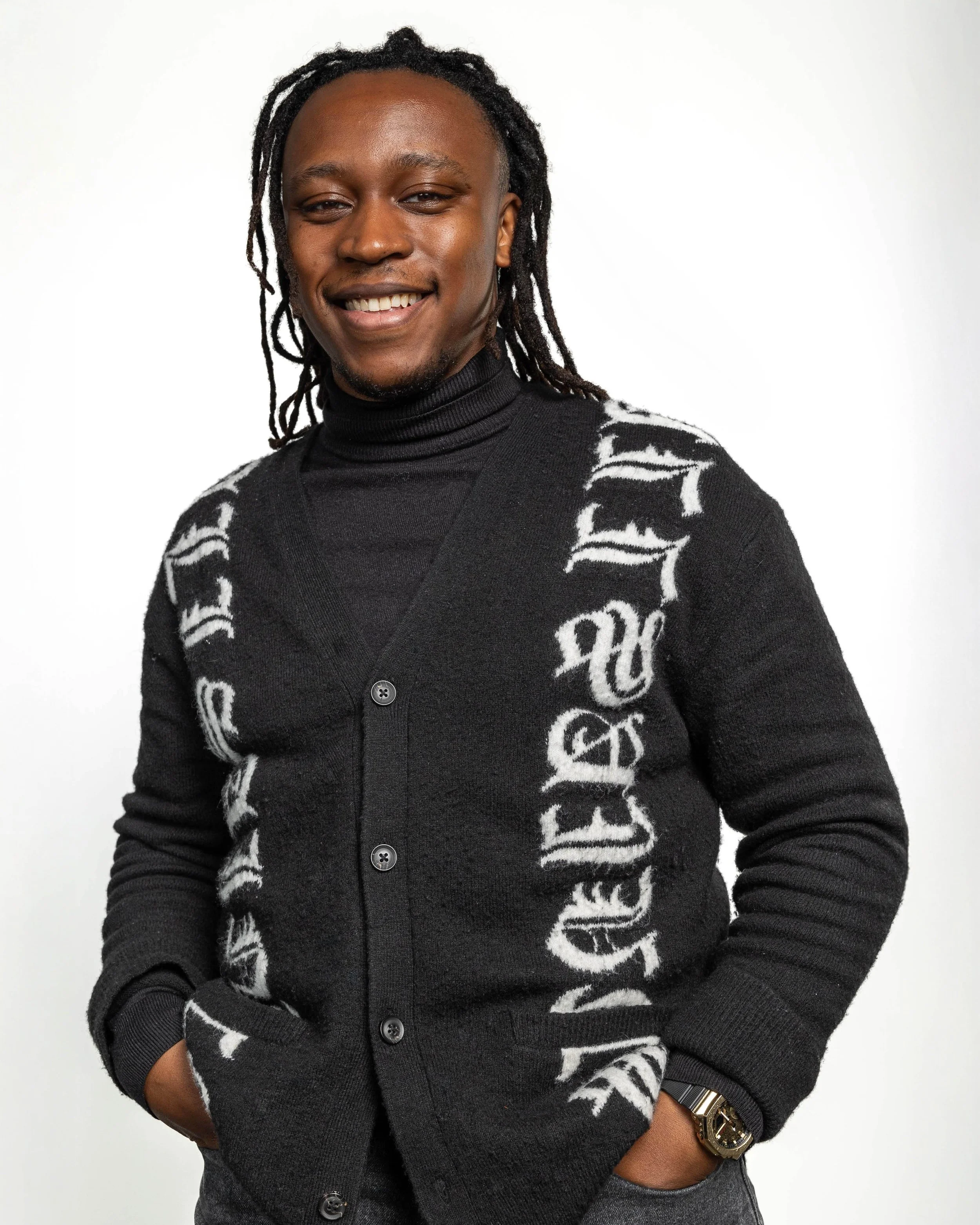 A young man with dreadlocks smiling and wearing a black sweater with white text, standing against a white background.