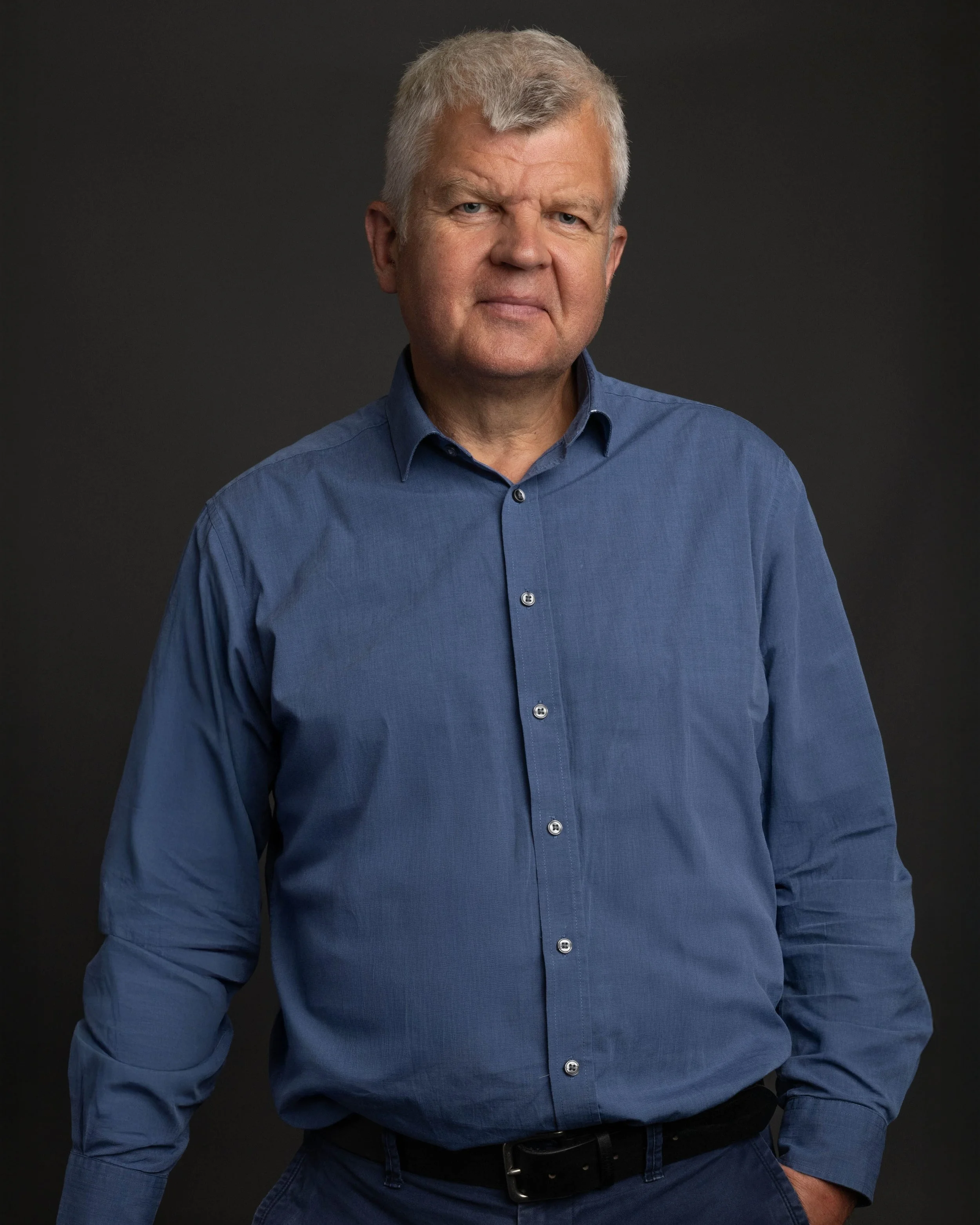 Portrait of a middle-aged man with gray hair wearing a blue dress shirt against a dark background.